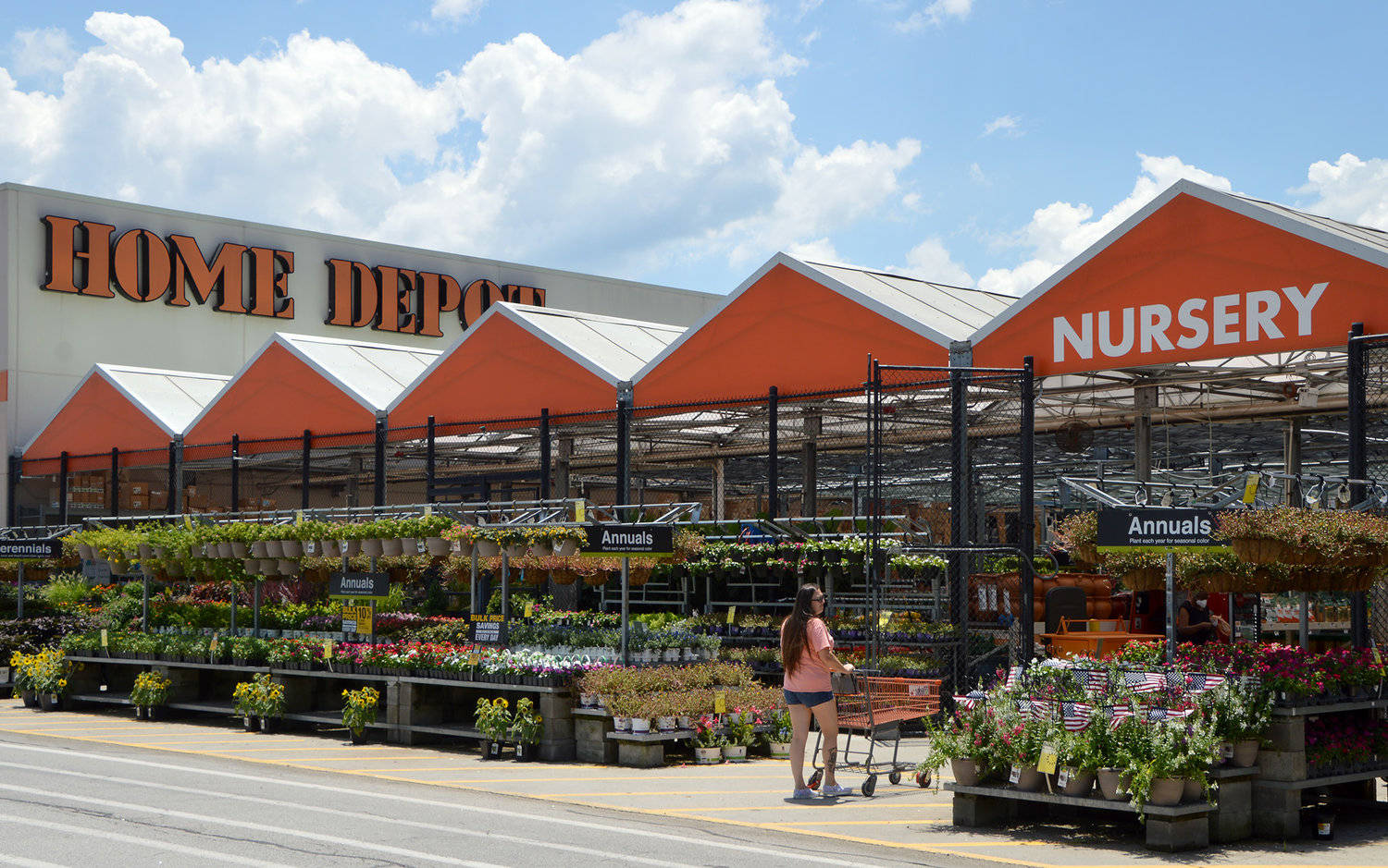 Home Depot 桌布 壁紙 — 在陽光明媚的日子裡，一名女性推著購物車穿過充滿色彩繽紛一年生植物的 Home Depot 苗圃，上方是鮮亮的橙色遮陽篷。