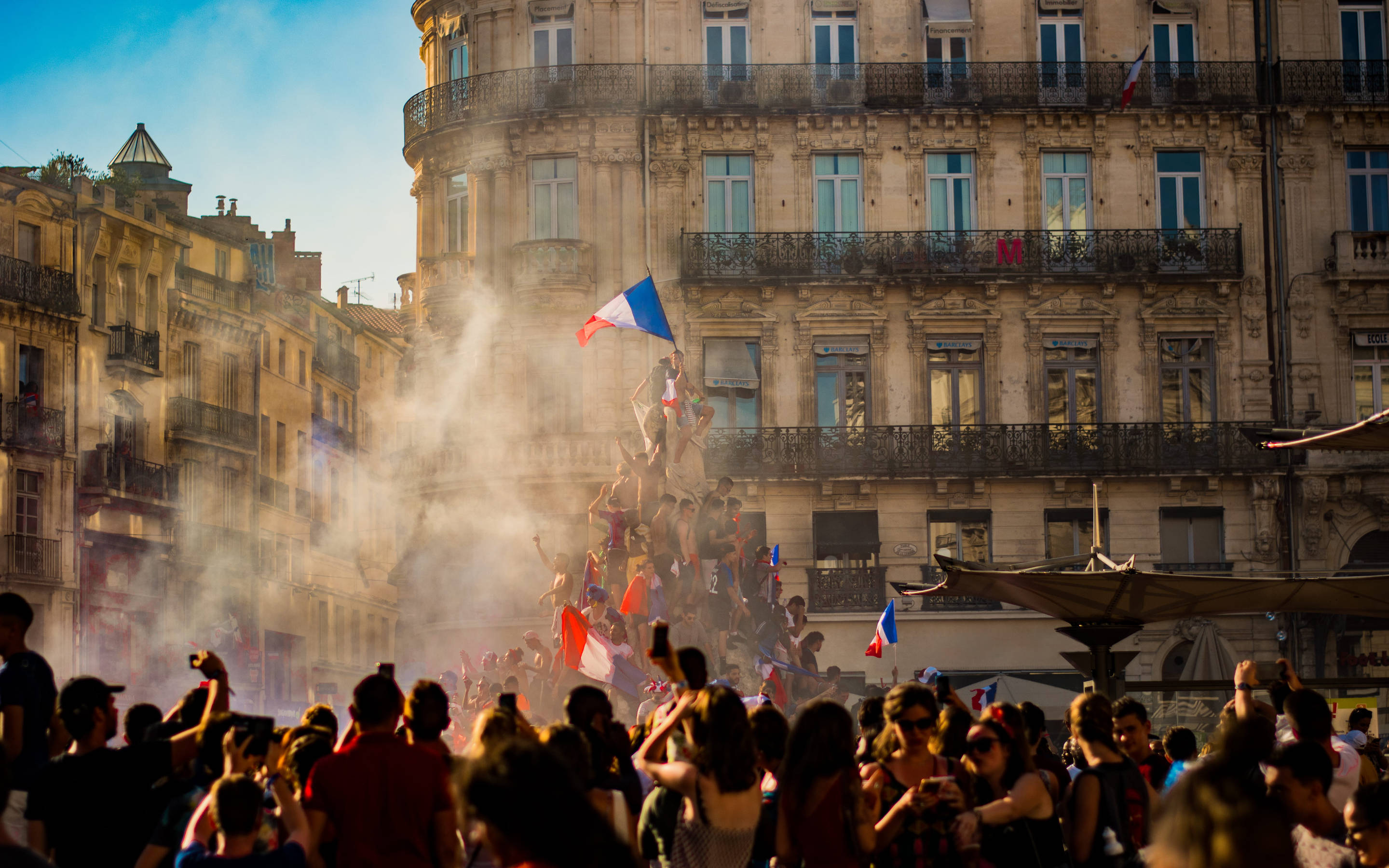 France Flag 桌布 壁紙 — 在陽光明媚的歐洲廣場上，歡呼的人群在歷史建築和煙霧中揮舞法國國旗慶祝。