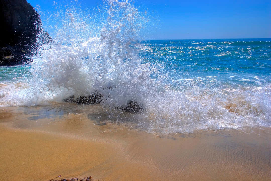 Beach Waves 桌布 壁紙 — 在陽光明媚的海灘上，一股強勁的海浪拍打著深色岩石，將耀眼的白色泡沫噴灑到空中。