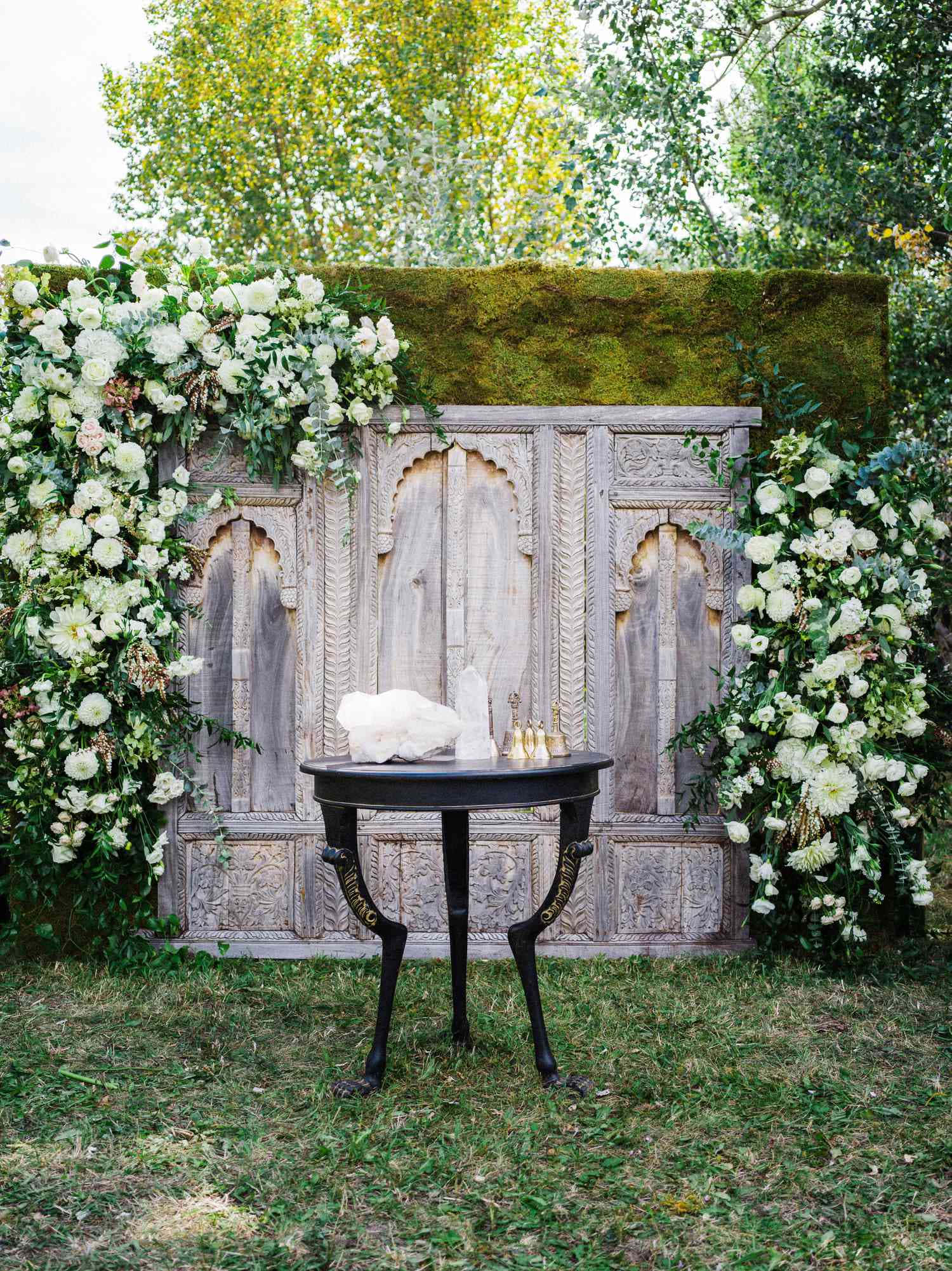 Garden Wedding 桌布 壁紙 — 在陽光明媚的花園中，一張黑色華麗桌子在長滿苔蘚的雕刻木屏風前放置著水晶和白色花朵。