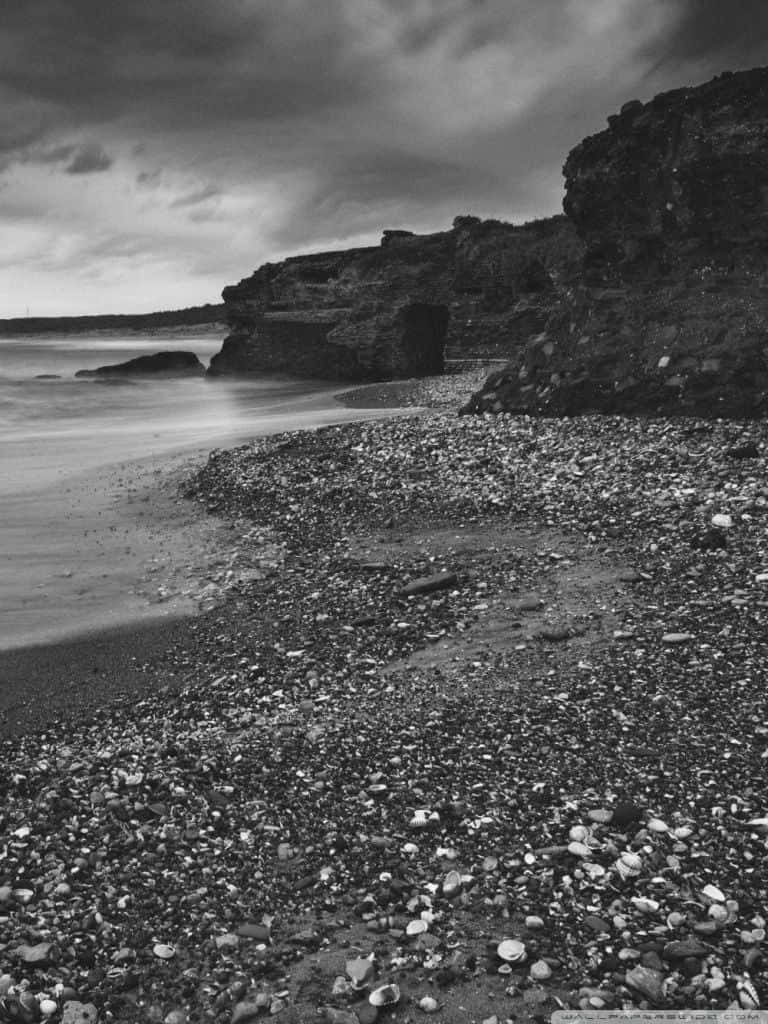 Black And White Beach 桌布 壁紙 — 在風暴天空下，卵石海灘上波浪模糊成霧氣，拍打崎嶇懸崖的單色長曝光照片。