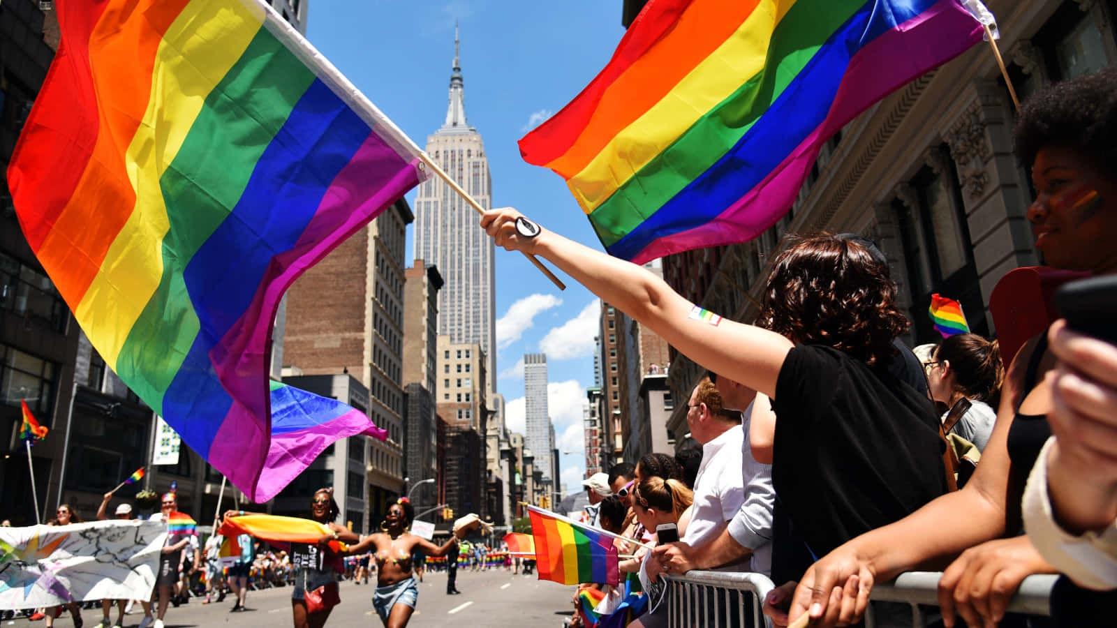 LGBT 桌布 壁紙 — 在驕傲遊行期間，鮮豔的彩虹旗高高飄揚在繁忙的城市街道上方，背景是帝國大廈。