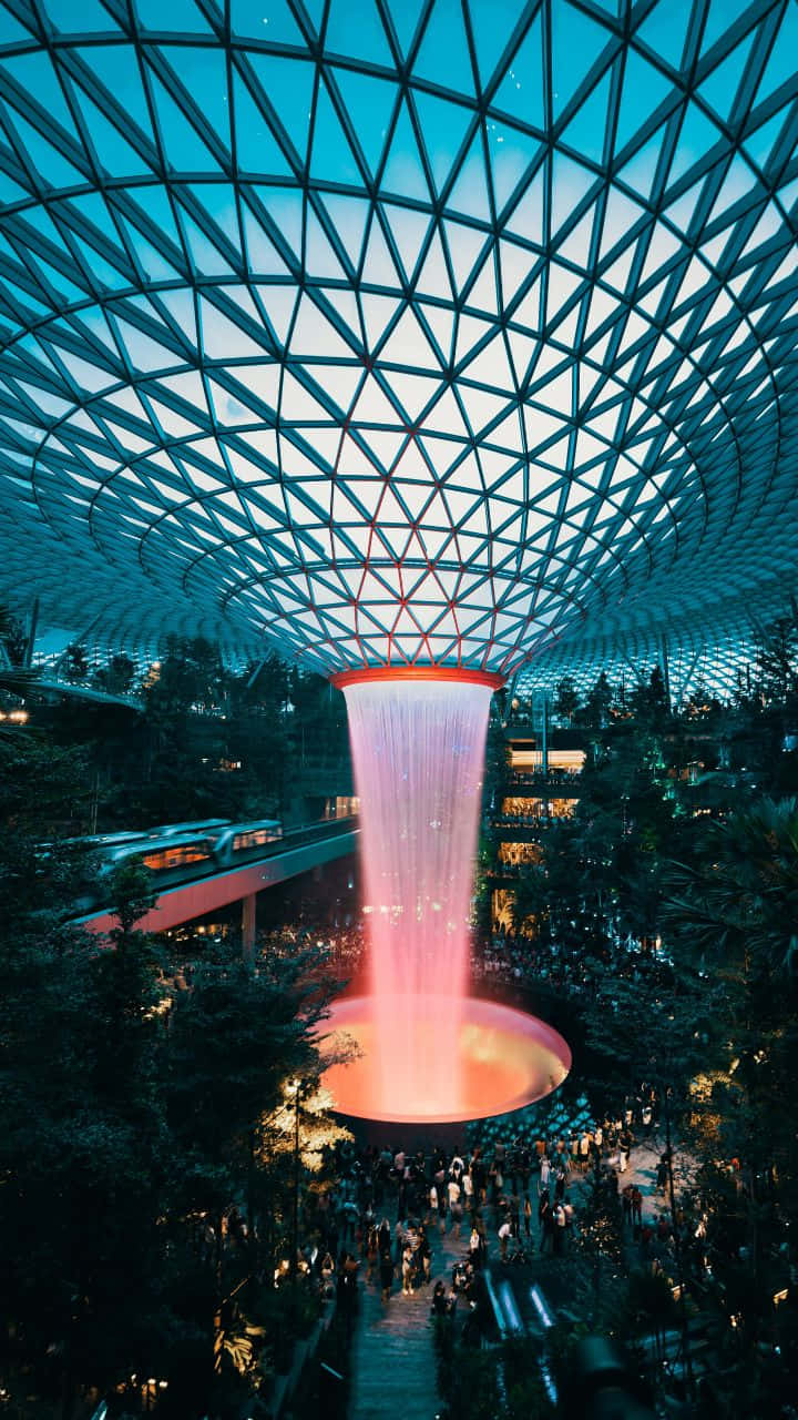 Singapore 桌布 壁紙 — 在黃昏時分，一個發光的粉紅色瀑布從幾何玻璃圓頂傾瀉而下，落在繁忙的室內花園上方。
