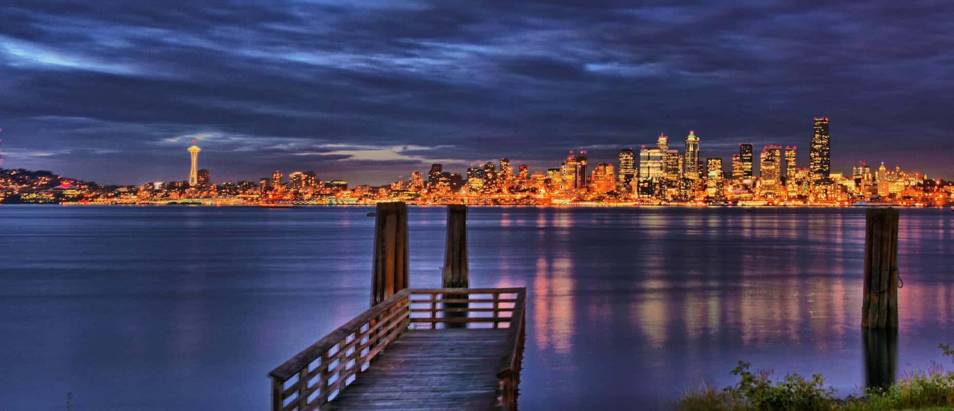 Seattle At Night 桌布 壁紙 — 在黃昏時分，一條木製碼頭延伸至平靜的水面，映照出在憂鬱藍天下閃爍的西雅圖天際線和太空針塔。