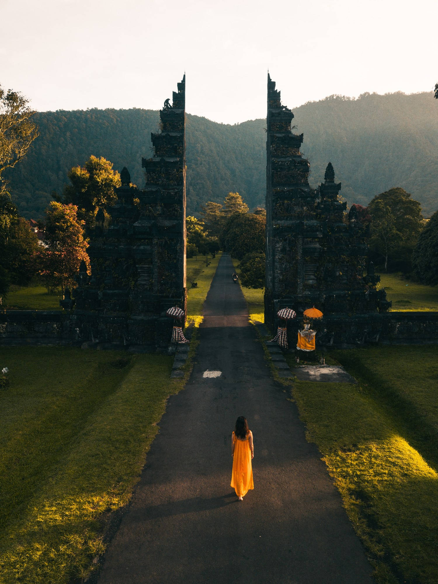Travel And Tourism 桌布 壁紙 — 在黃金時刻，一位身穿飄逸黃色連衣裙的孤獨女子沿著石徑走向古老的峇里島寺廟大門，四周是鬱鬱蔥蔥的青山。