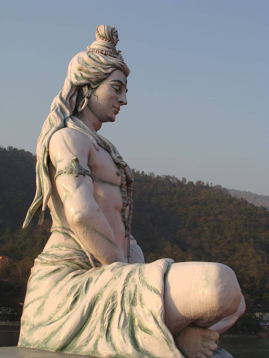 bholenath 桌布 壁紙 — 在黃金時刻，一尊寧靜的濕婆神石像以冥想姿勢俯瞰著河流，背景是森林覆蓋的山丘。