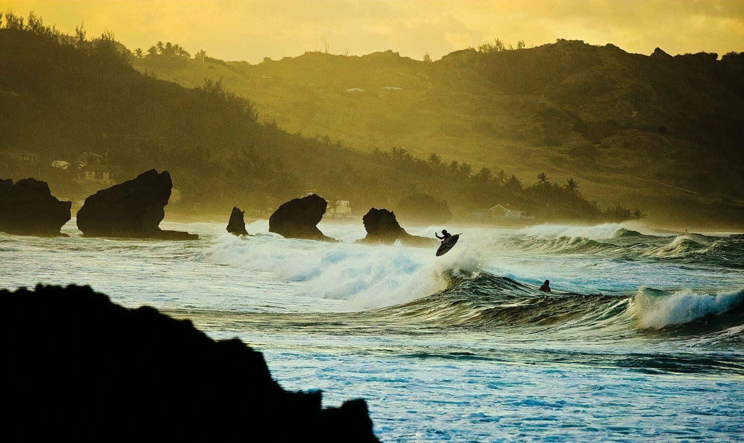 surfing 桌布 壁紙 — 在黃金時段，一名衝浪者乘著破碎的海浪，背景是岩石海岸和棕櫚樹的剪影。