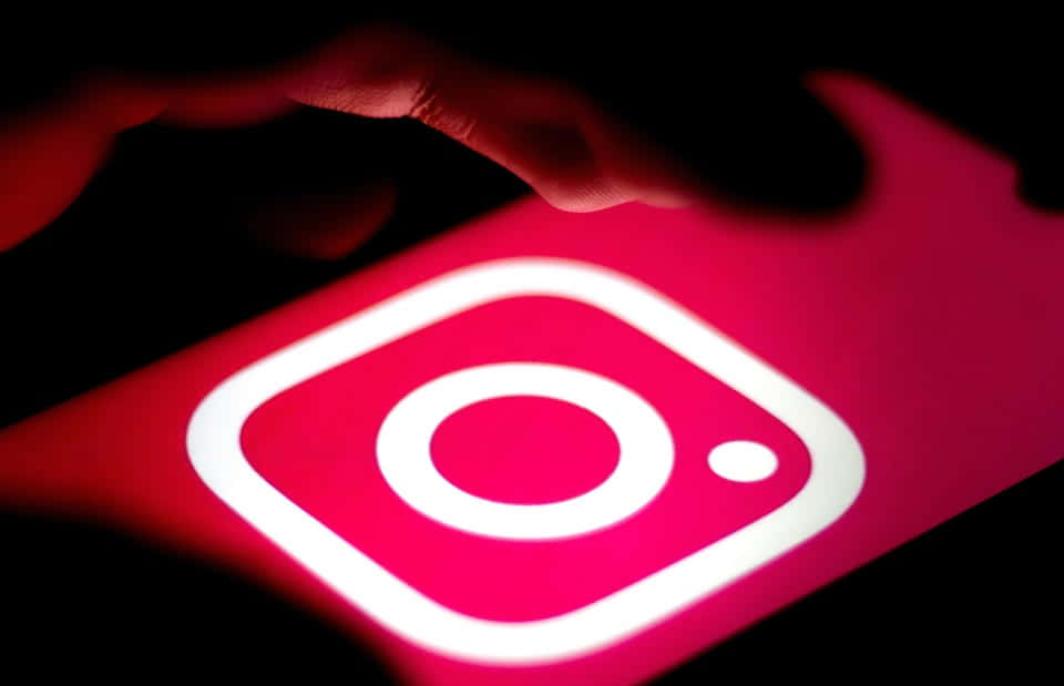 Logo Design 桌布 壁紙 — 在黑暗中，智慧型手機螢幕上有一個發光的紅色Instagram標誌，手指懸浮在其上方。