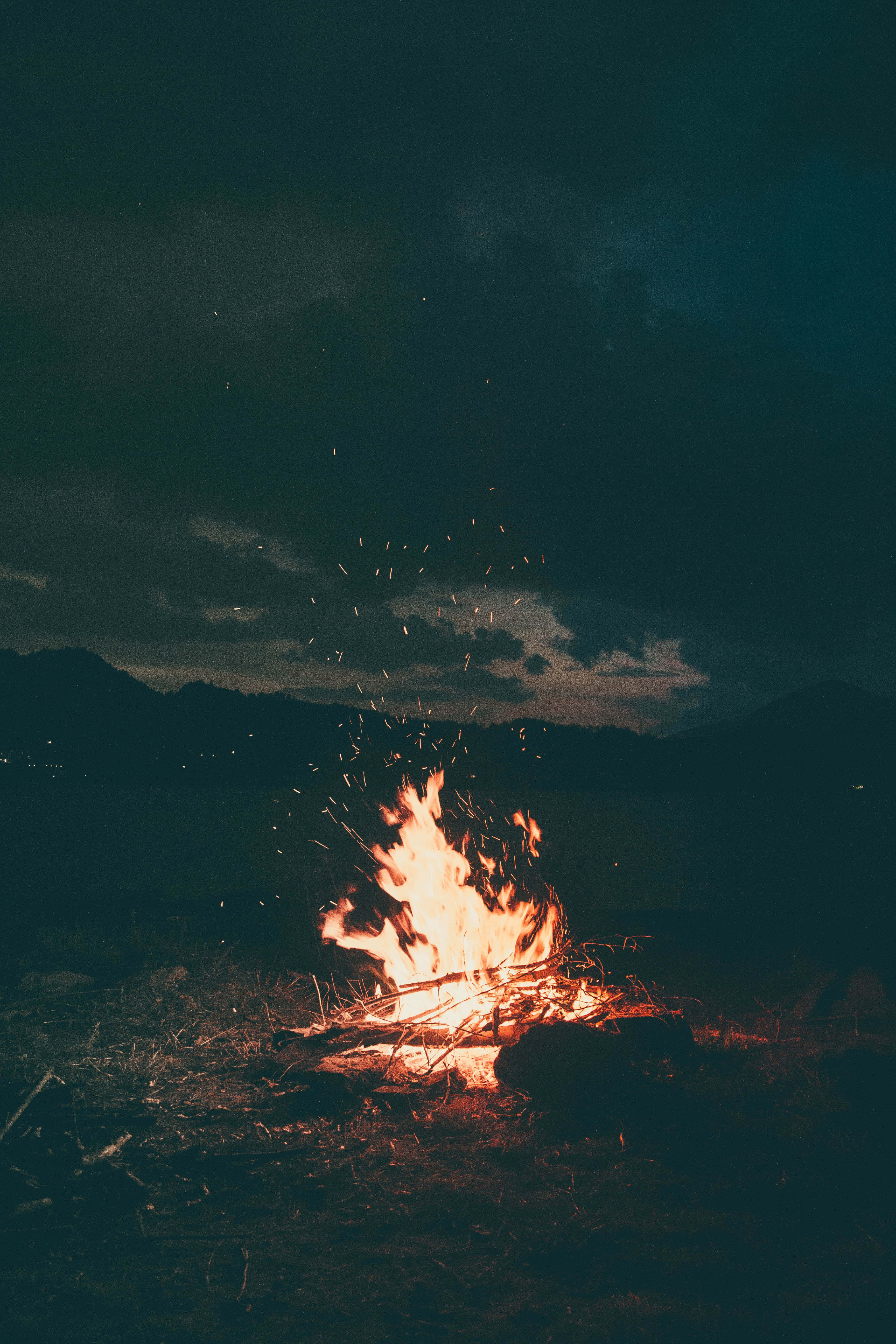Bonfire 桌布 壁紙 — 在黑暗多雲的天空下，一個充滿活力的營火劈啪作響，發光的餘燼在夜間荒野環境中向上飛舞。
