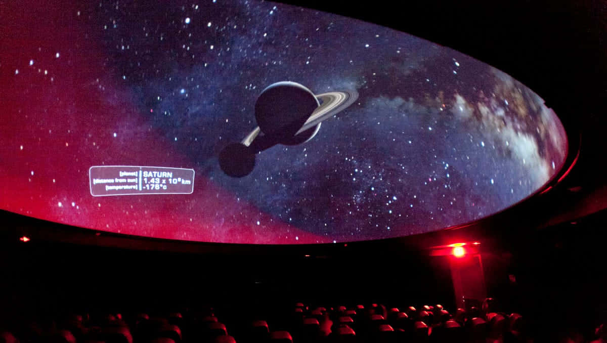 Planetarium 桌布 壁紙 — 在黑暗的劇院中，彎曲的圓頂螢幕顯示帶有光環的土星，背景是繁星點點的宇宙，並有資料疊加。