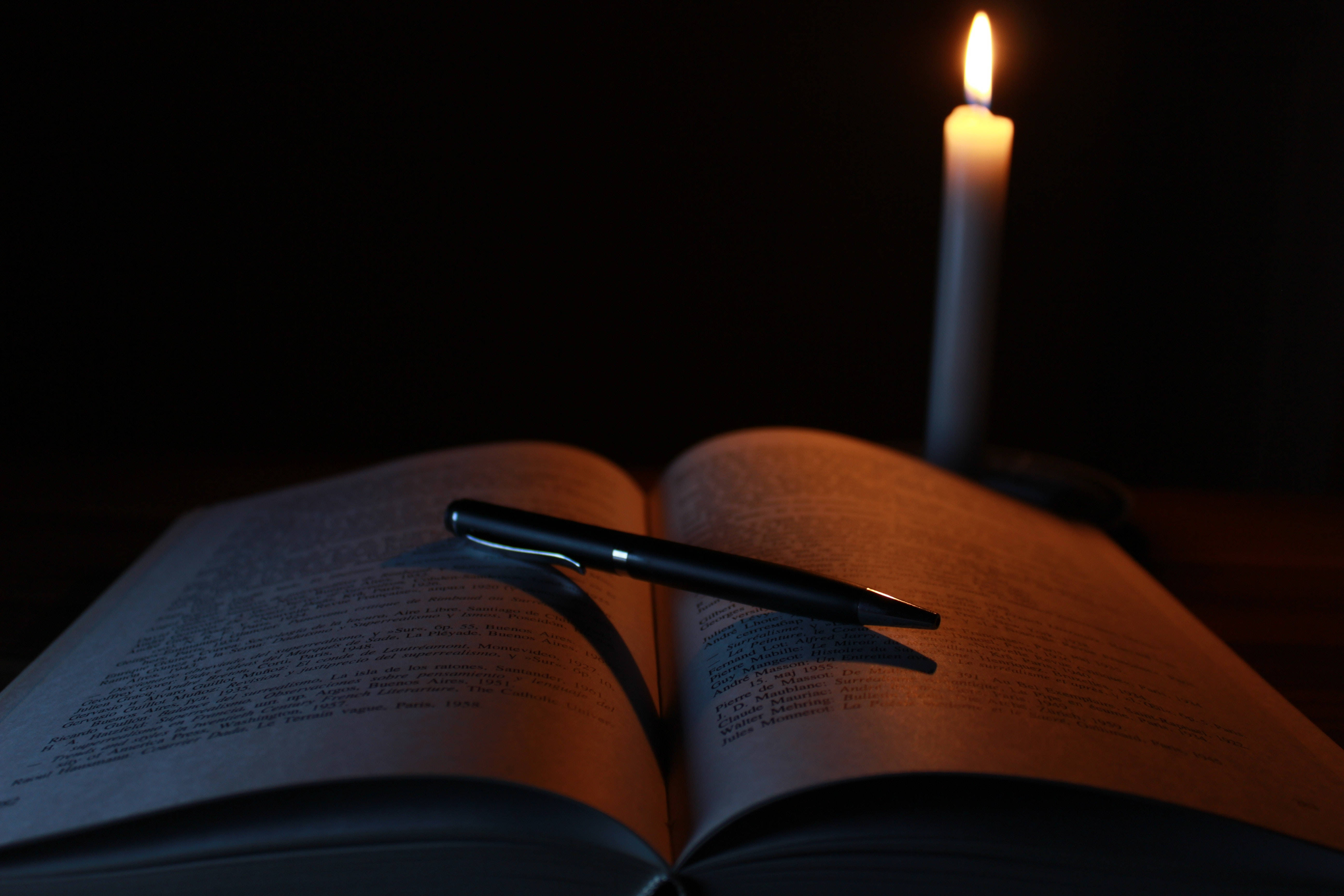 讀書動力 桌布 壁紙 — 在黑暗的房間中，一支蠟燭將溫暖的光芒投射在一本打開的書上，一支筆擱在書頁上。