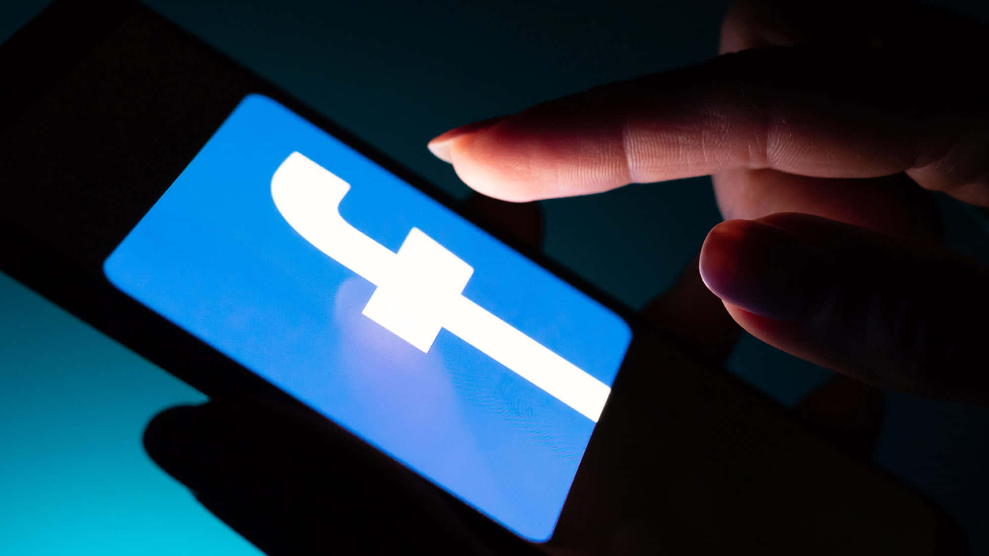 Facebook 桌布 壁紙 — 在黑暗的房間中，一隻手點擊智慧型手機螢幕上發光的藍色Facebook標誌。