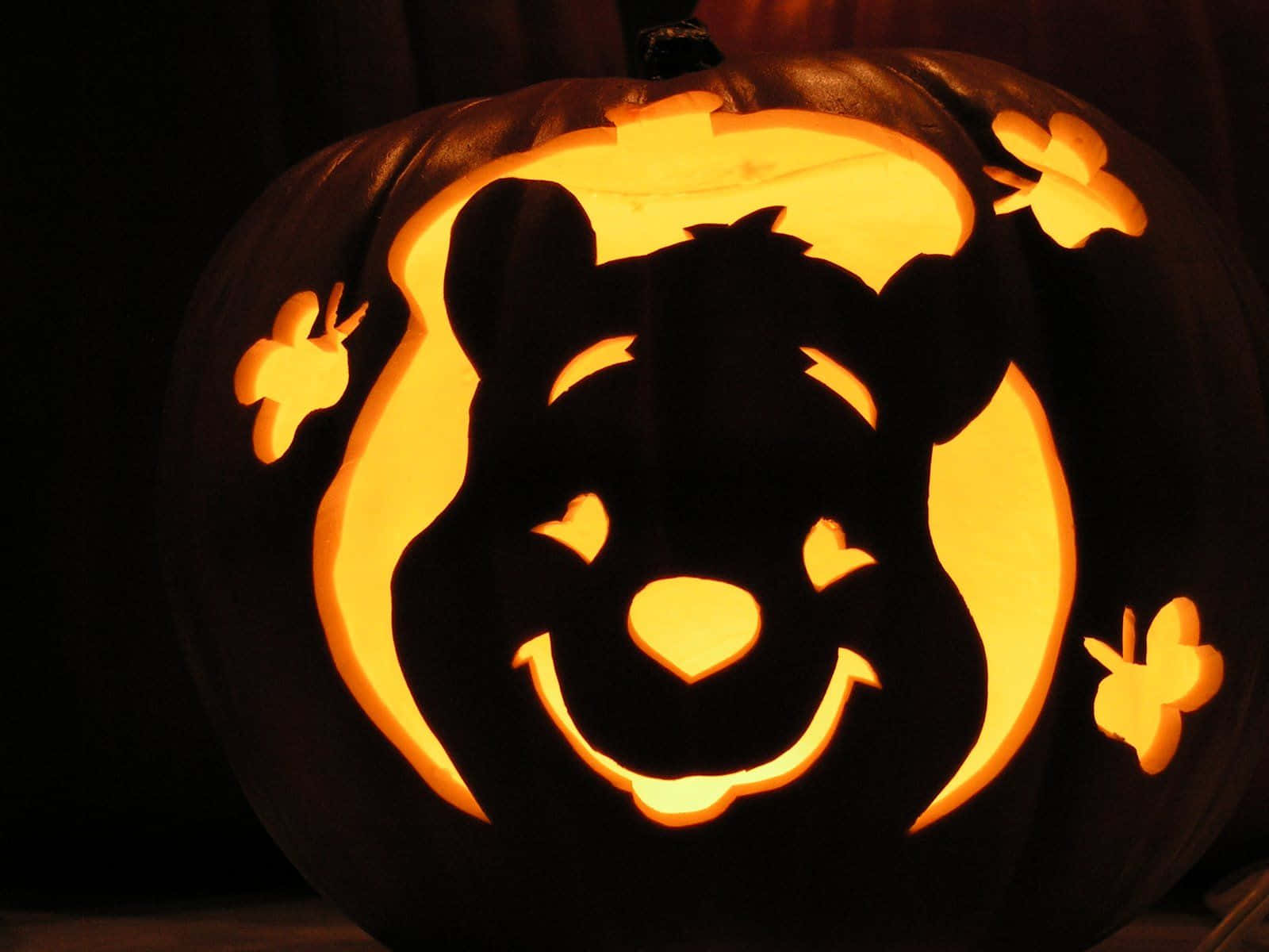 Winnie The Pooh Halloween 桌布 壁紙 — 在黑暗背景下，一個發光的南瓜燈雕刻著微笑的小熊維尼臉和蝴蝶裝飾。