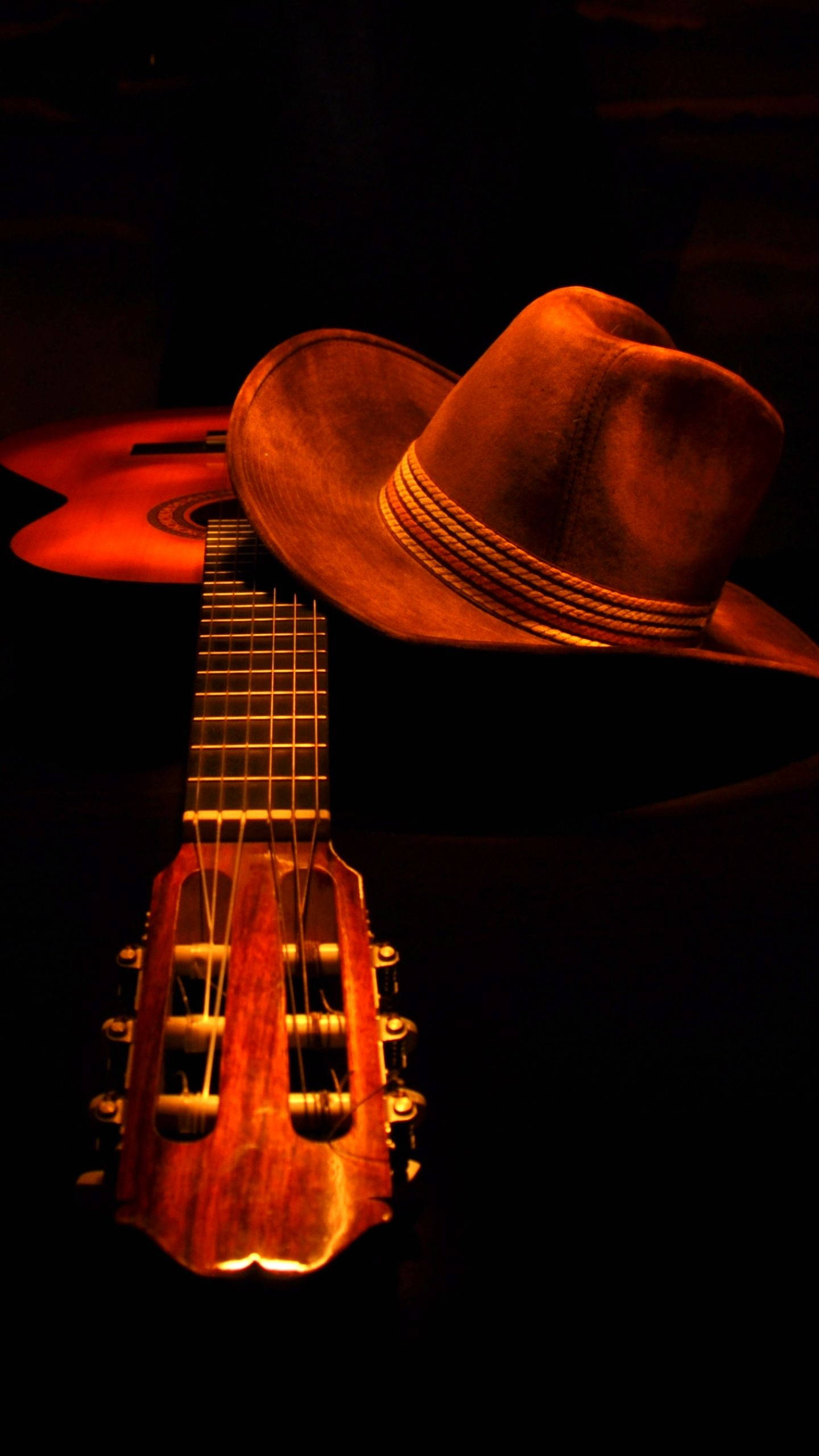 Guitar Aesthetic 桌布 壁紙 — 在黑暗背景下，一盞溫暖照明的原聲吉他，其琴頸上放著一頂牛仔帽。