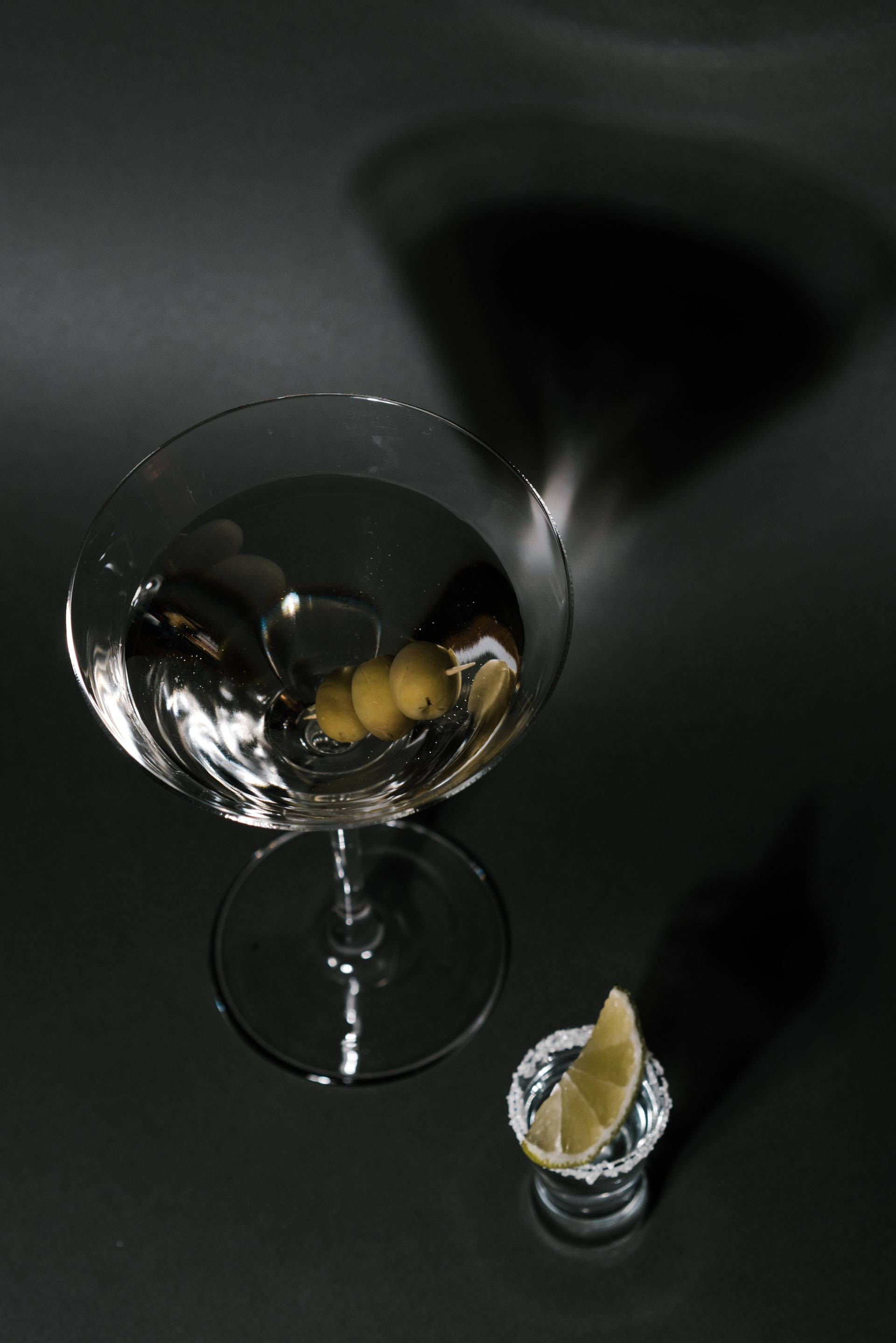 martini 桌布 壁紙 — 在黑暗表面上，一個馬丁尼杯裝有橄欖和萊姆角，在戲劇性燈光下。