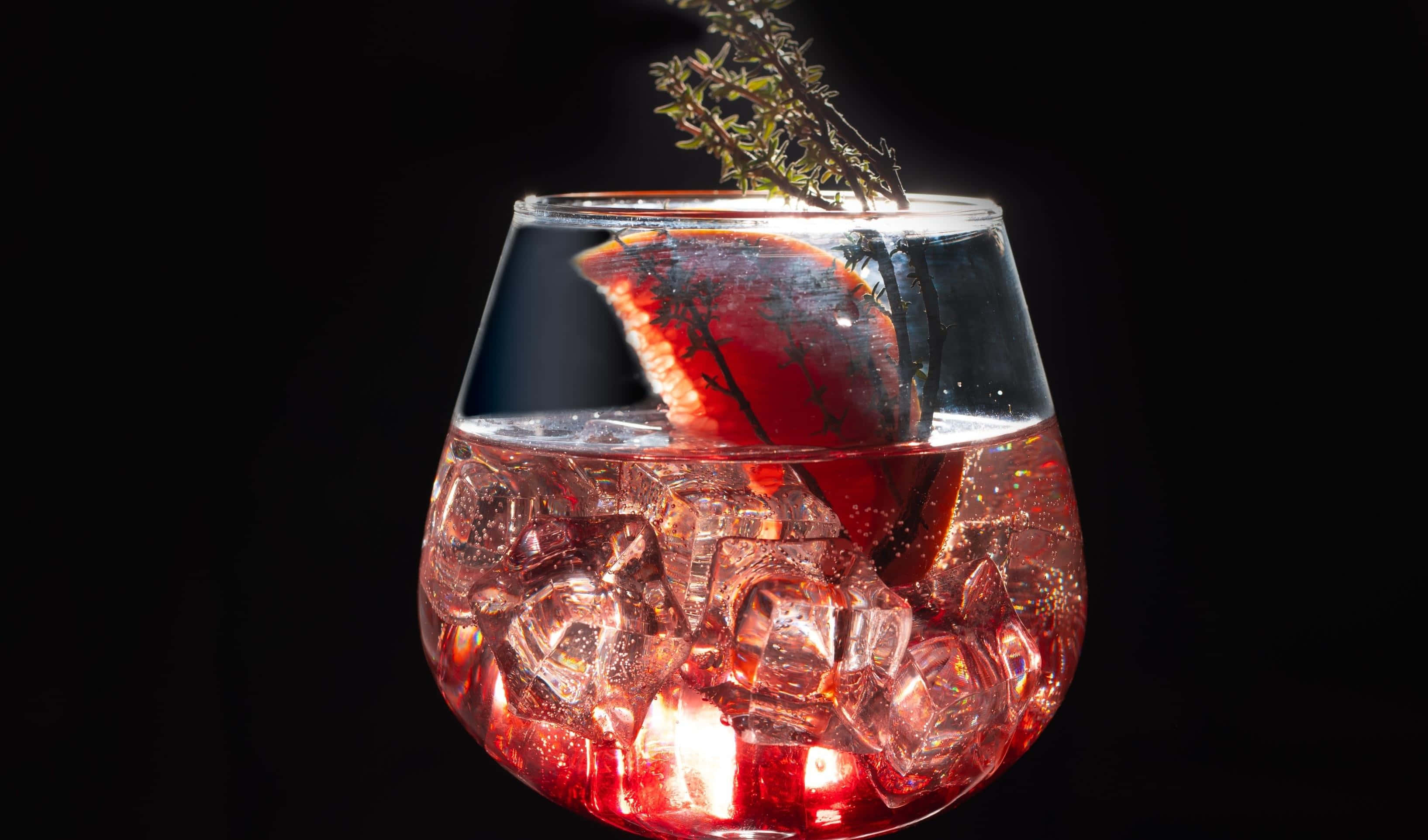 Halloween Cocktails 桌布 壁紙 — 在黑色背景前，一杯發光的紅色雞尾酒，裝滿冰塊，並以柑橘片和香草枝裝飾。