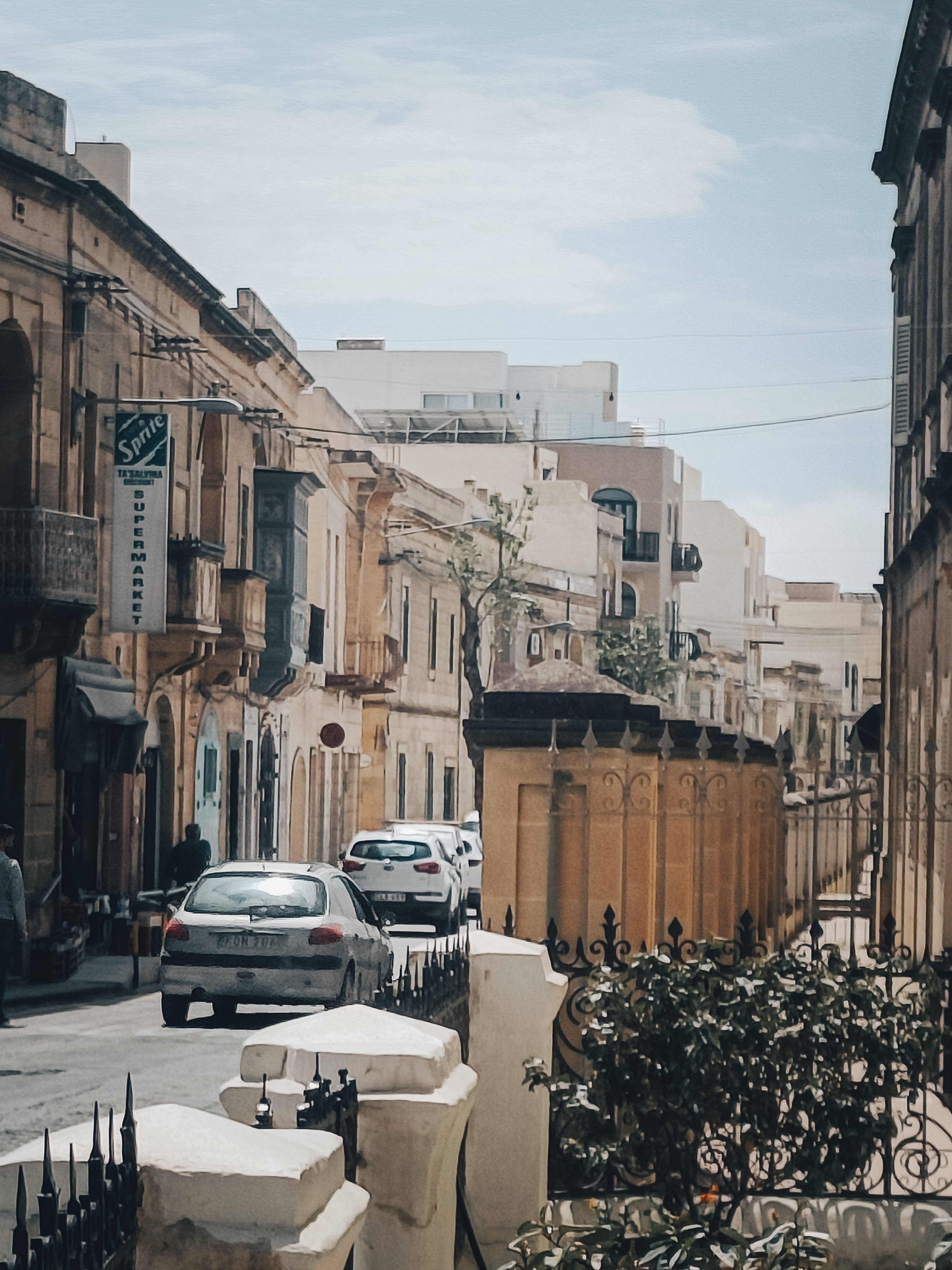 Malta 桌布 壁紙 — 地中海小鎮一條狹窄的陽光街道，有粉彩建築、汽車和華麗的黑色鐵欄杆。