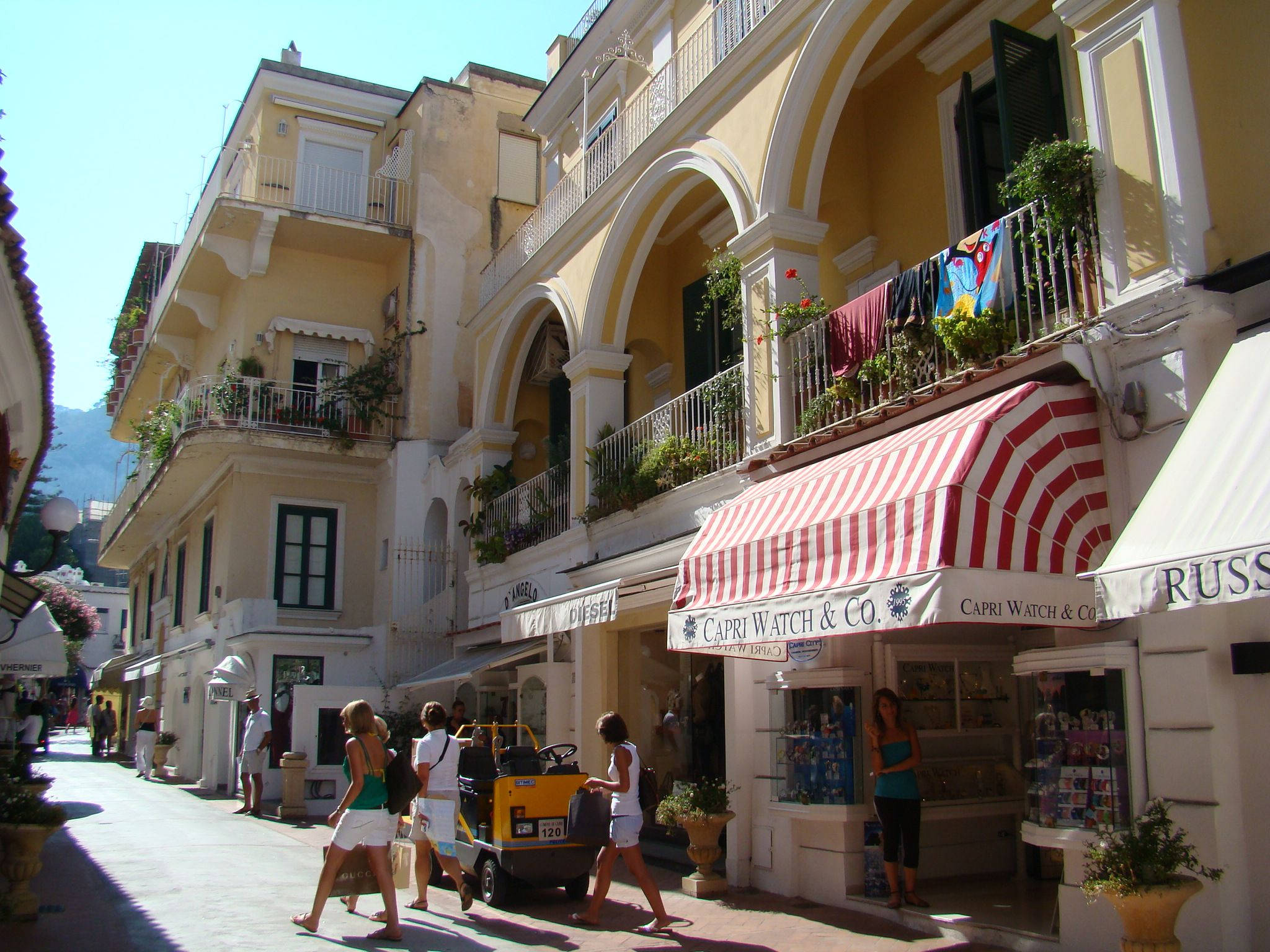 Capri 桌布 壁紙 — 地中海小鎮的陽光鵝卵石街道，粉彩建築、種滿植物的陽台和條紋遮陽篷。