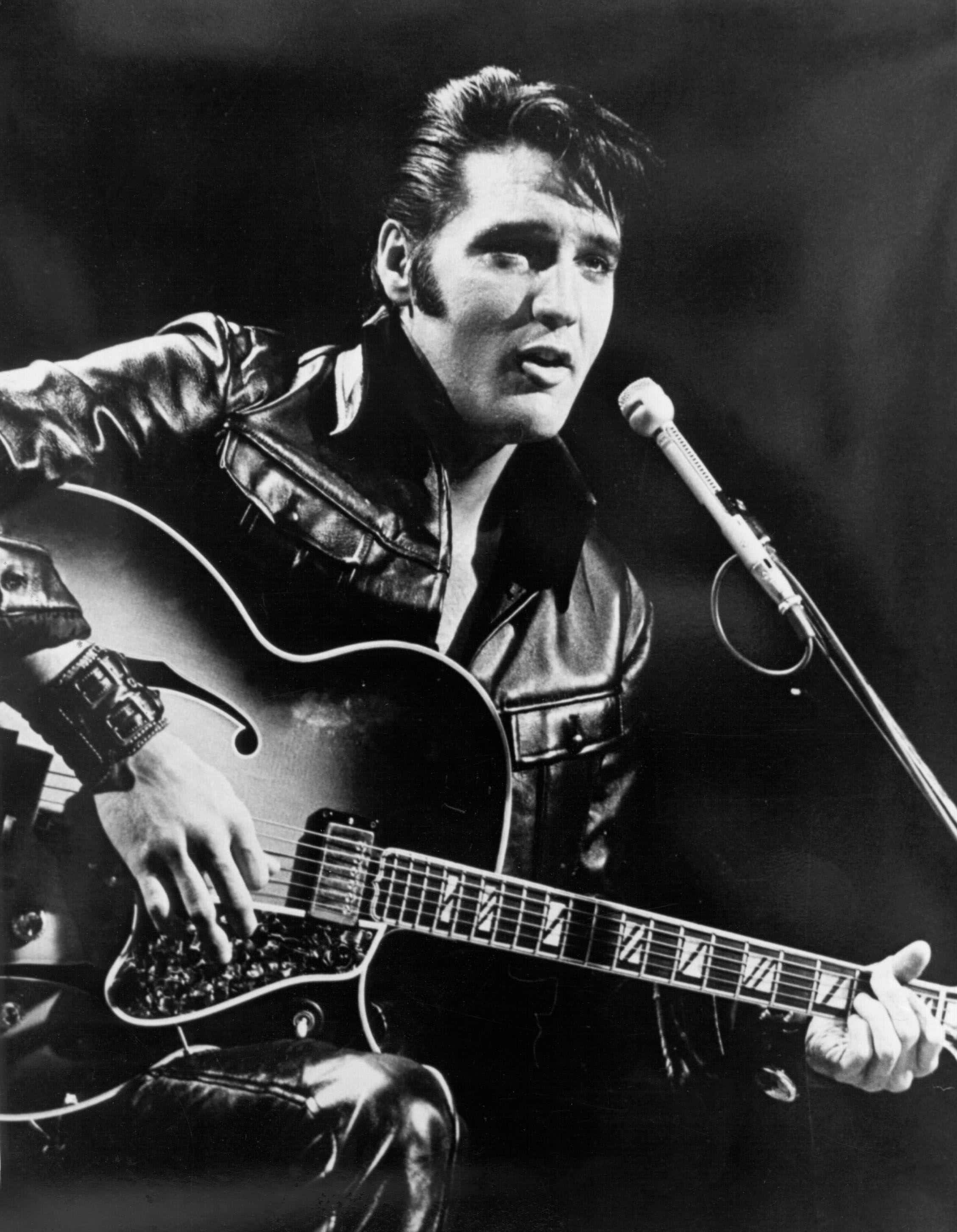 4K Elvis 桌布 壁紙 — 埃爾維斯·普里斯萊身穿皮夾克，手持電吉他，在戲劇性的舞台燈光下熱情表演。