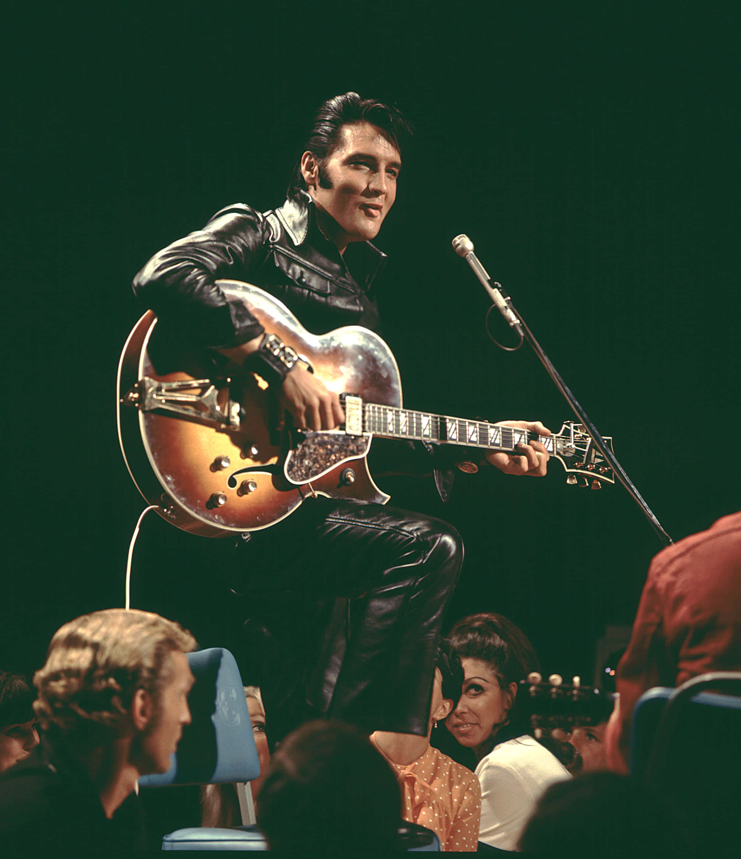 Elvis 桌布 壁紙 — 埃爾維斯·普雷斯利身穿黑色皮衣，手持旭日紋電吉他，在舞台燈光下為現場音樂會帶來震撼。