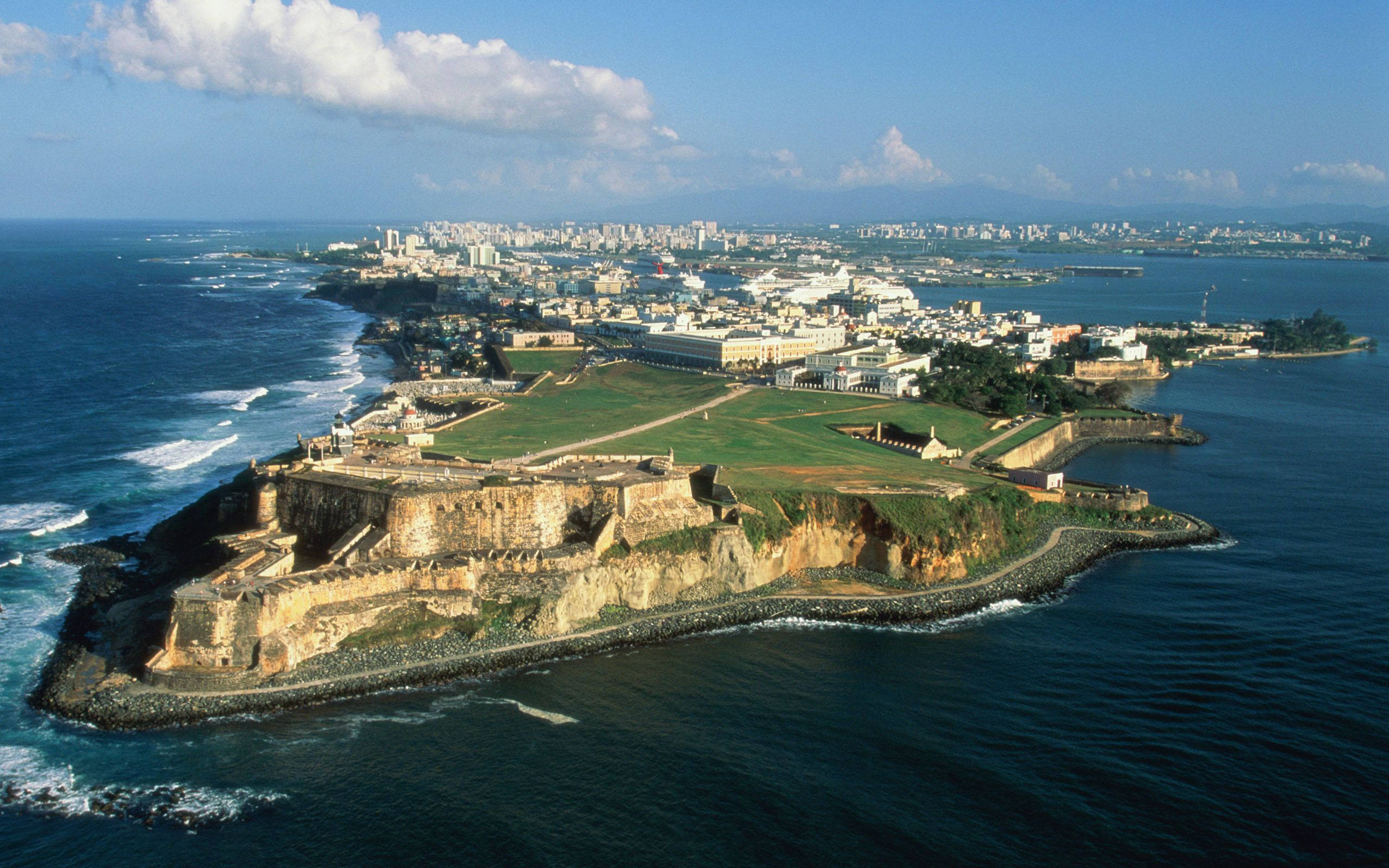 Puerto Rico 桌布 壁紙 — 埃爾莫羅要塞的空中景觀，坐落在岩石半島上，俯瞰著充滿活力的沿海城市和深藍色的海洋，天空部分多雲。