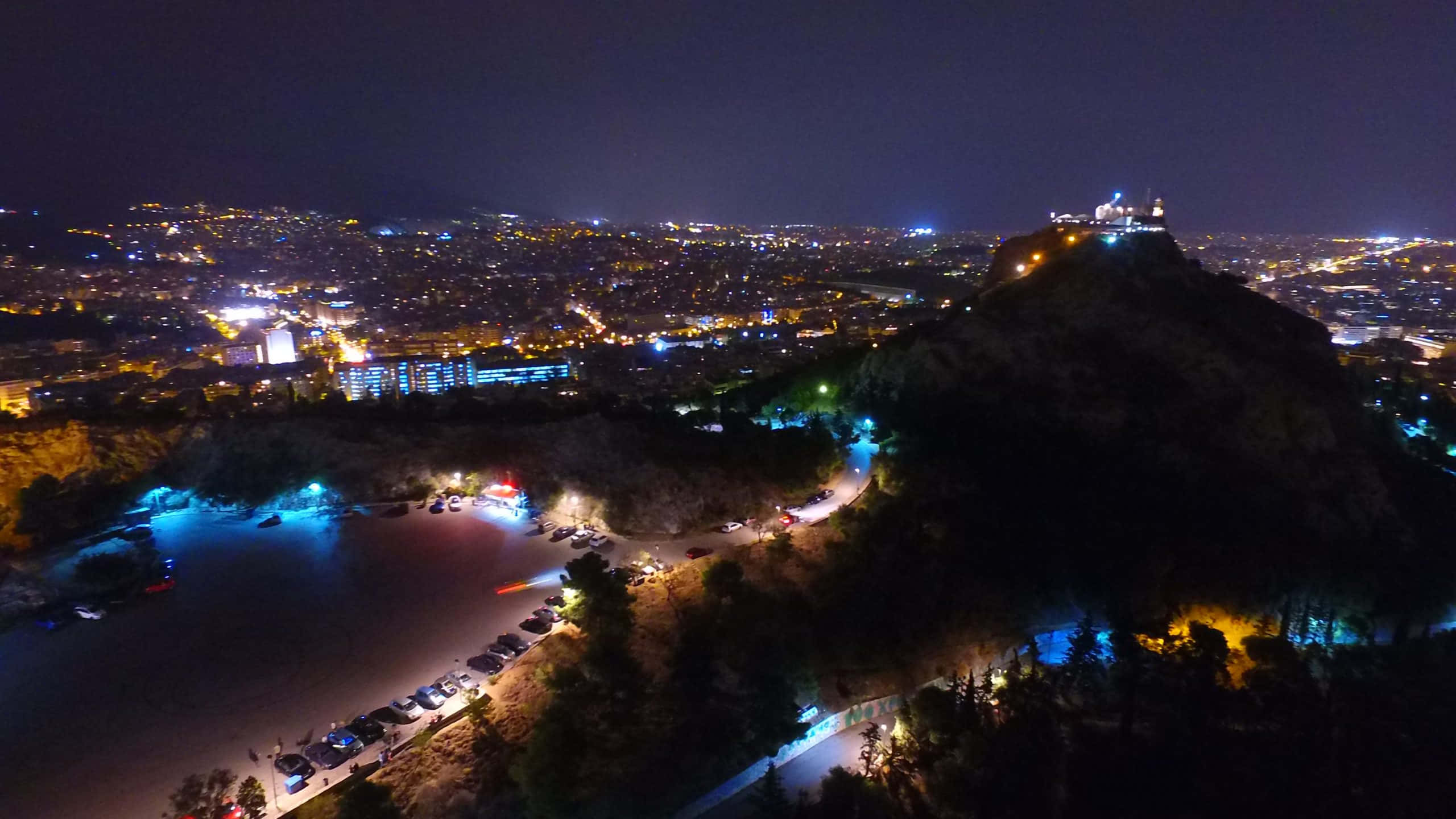 山景 桌布 壁紙 — 城市在燈火通明的山頂堡壘下方延伸的夜間鳥瞰圖，俯瞰著閃閃發光的水岸。