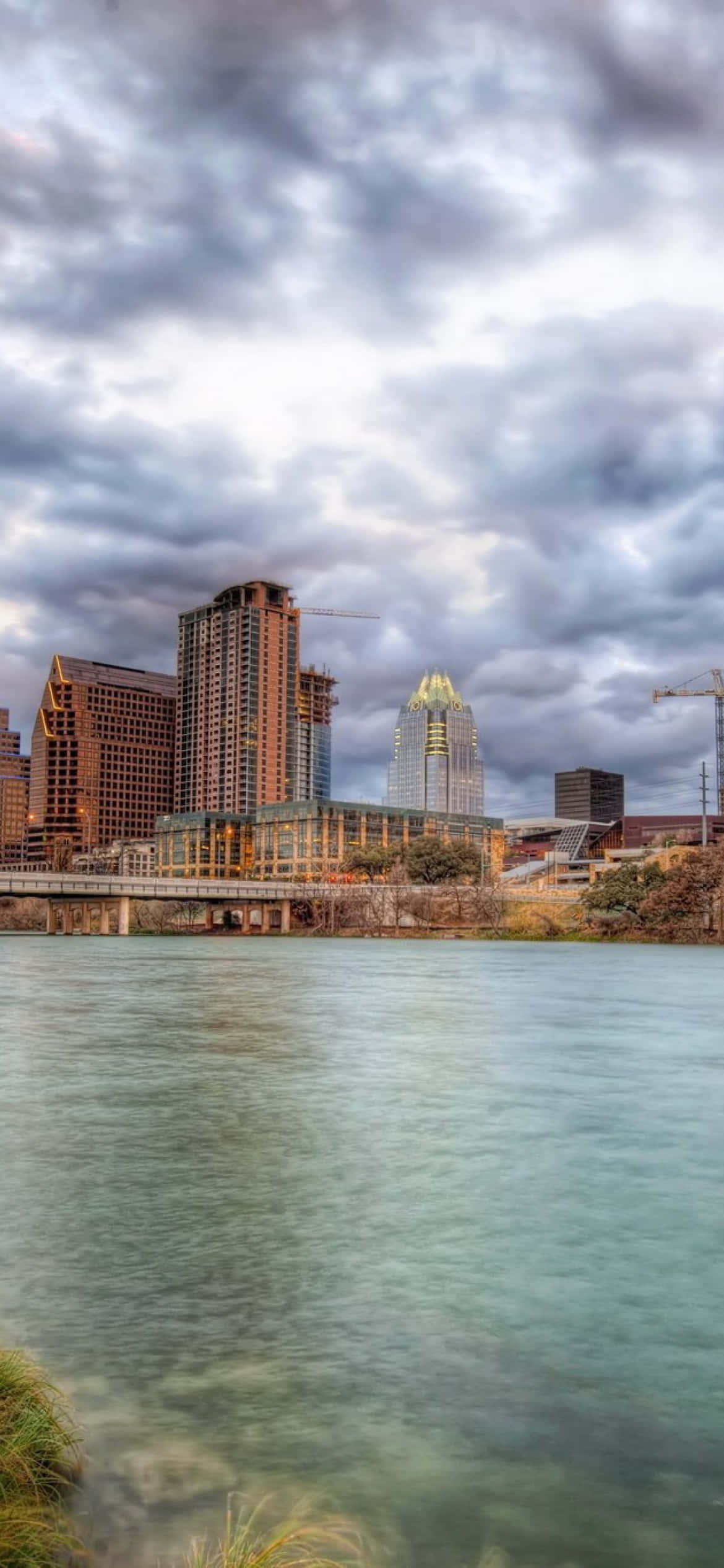 Texas iPhone 桌布 壁紙 — 城市天際線，現代摩天大樓和一座橋樑俯瞰著平靜的河流，天空烏雲密佈，氣勢磅礴。