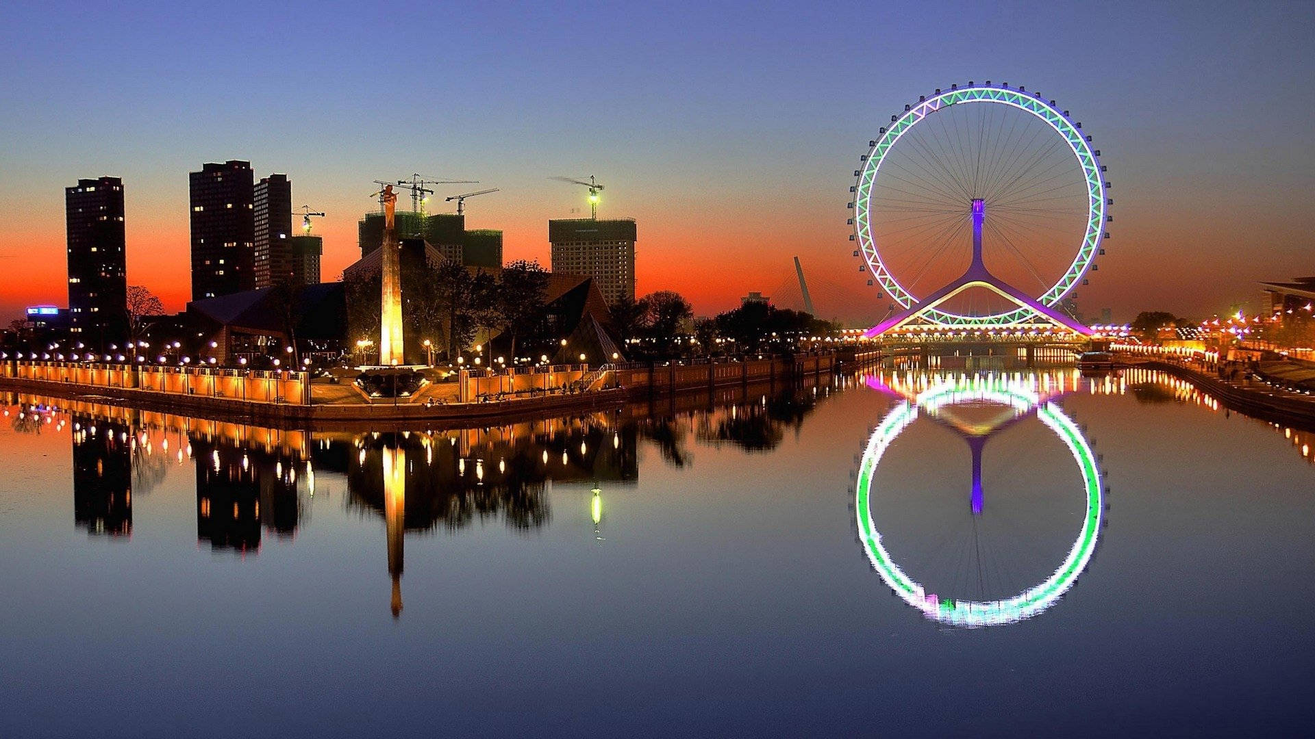 Tianjin 桌布 壁紙 — 城市天際線上的 vibrant 日落，發光的摩天輪倒映在平靜的水面上。