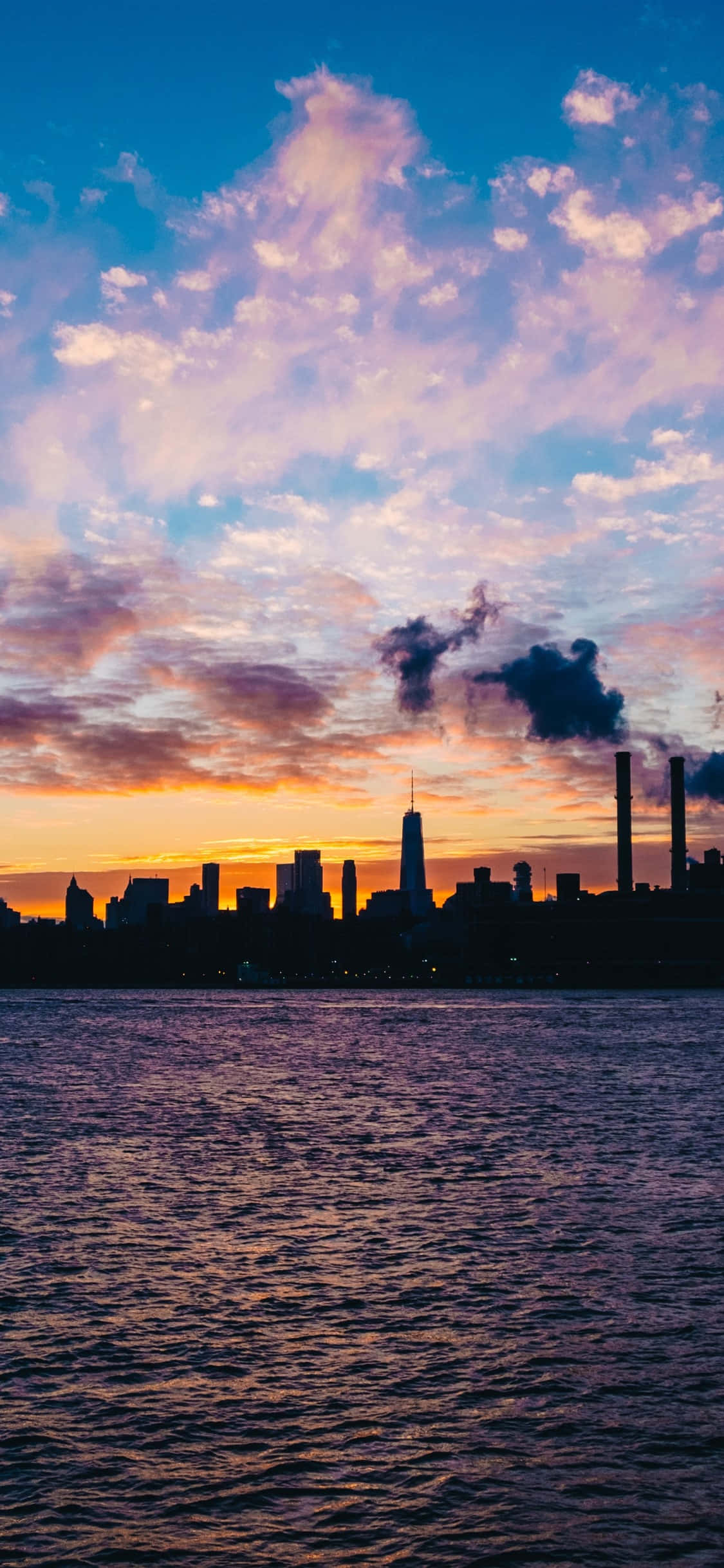 iPhone Xs New York 桌布 壁紙 — 城市天際線在充滿活力的日落天空下呈現剪影，粉紅色、橙色和藍色的雲彩倒映在漣漪的水面上。