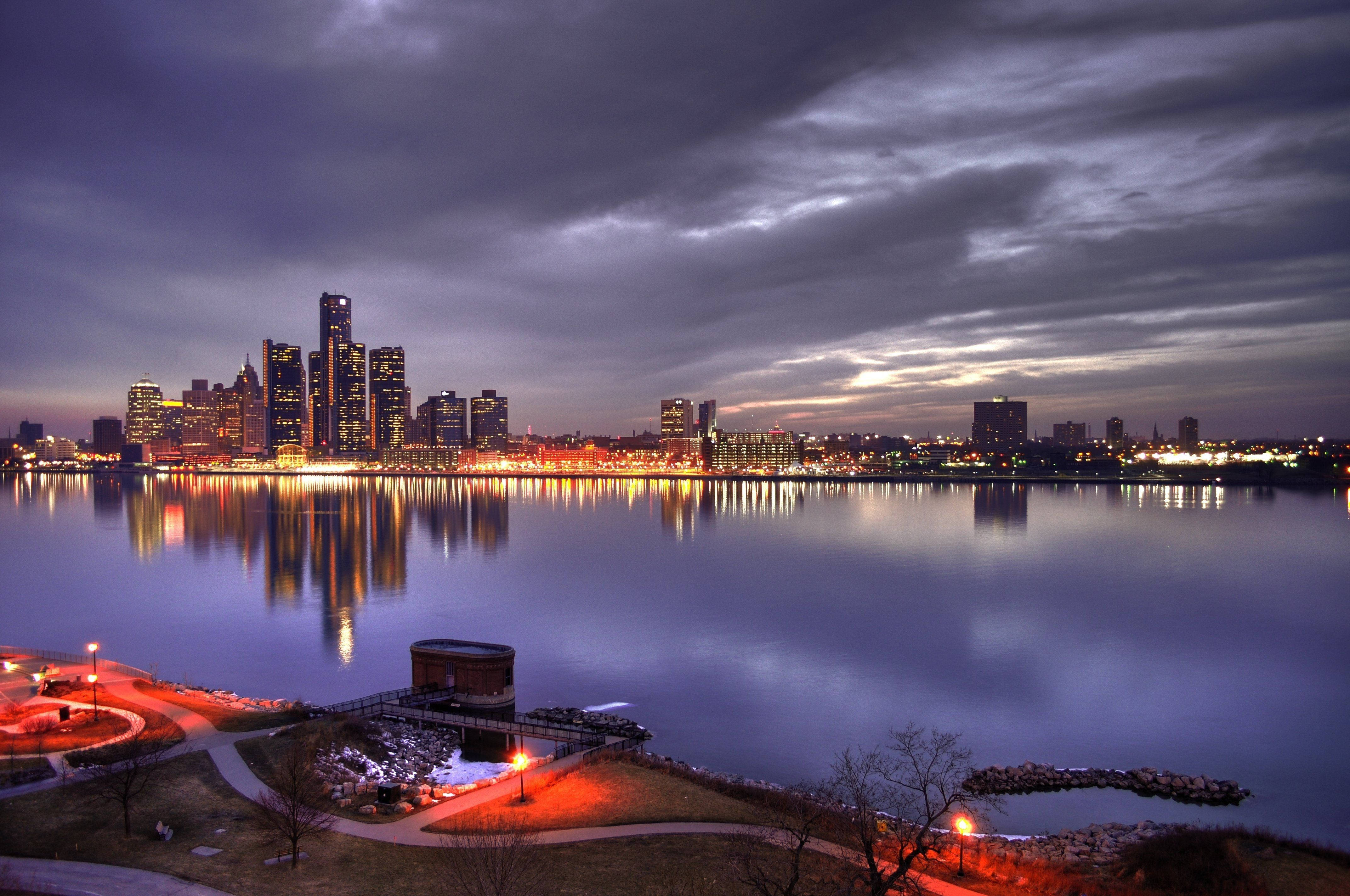 detroit 桌布 壁紙 — 城市天際線在暮色蒼茫的天空下閃爍著溫暖的燈光，倒映在平靜的水面上。