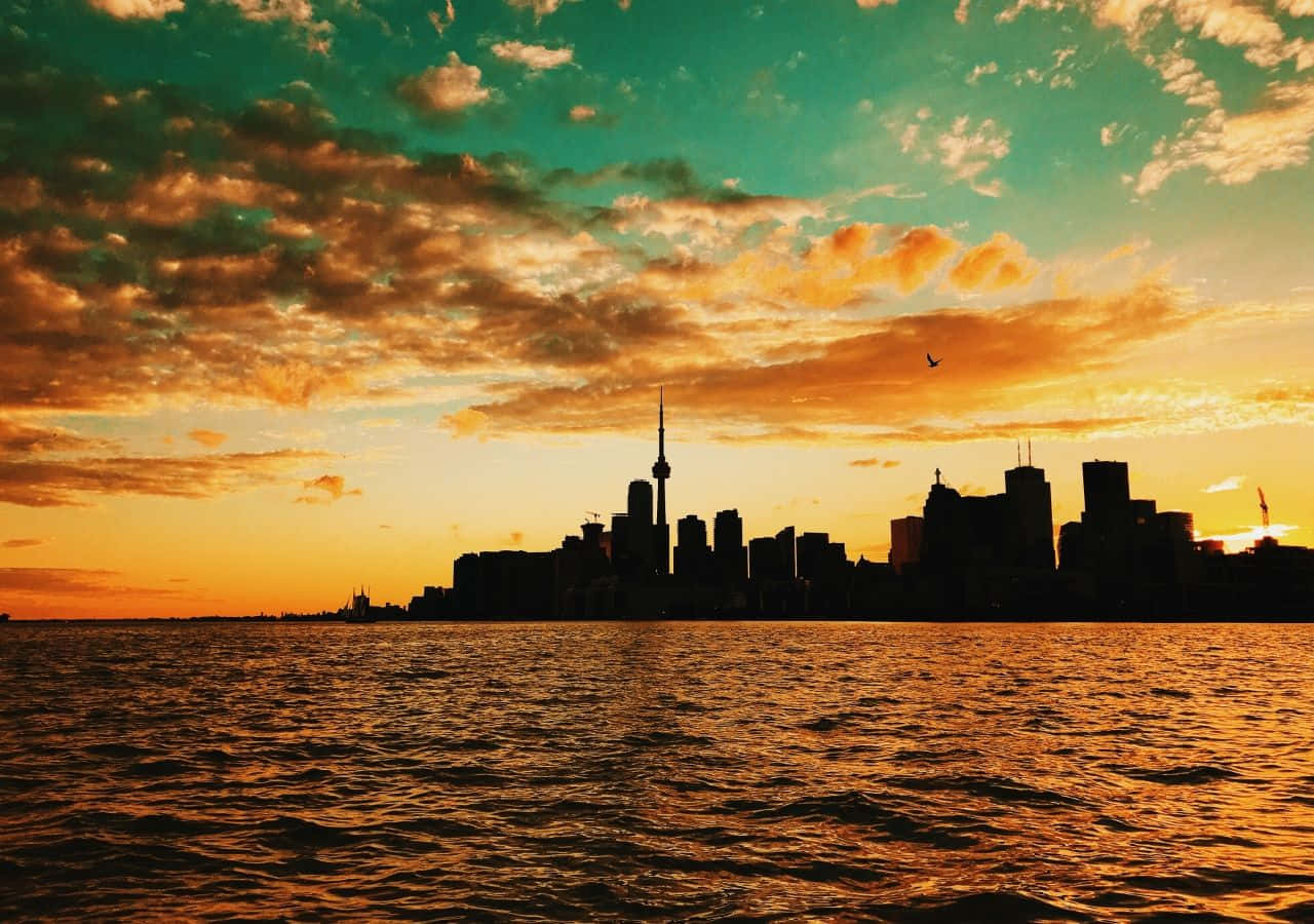 Toronto 桌布 壁紙 — 城市天際線在火熱的橙色和青綠色日落映襯下，倒映在漣漪的水面上。