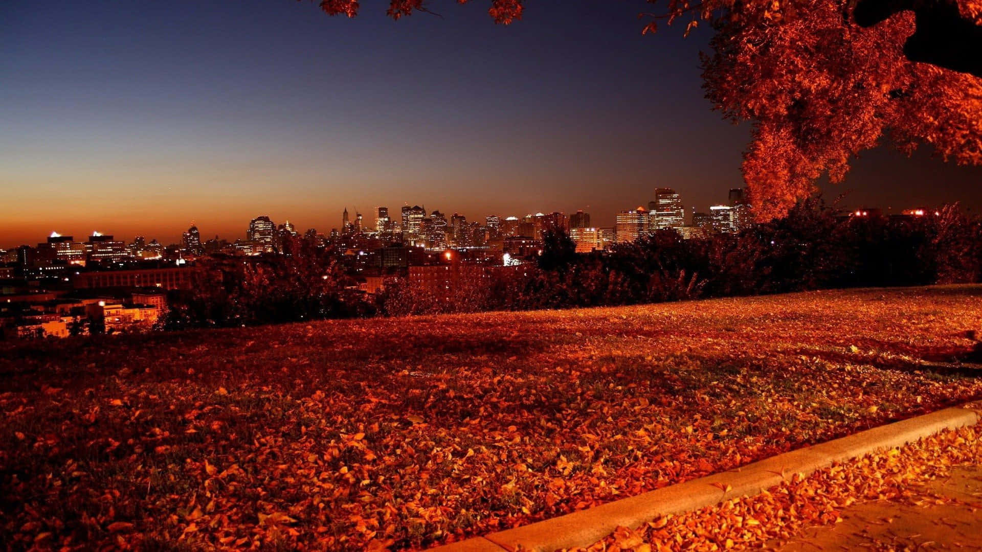 Fall City 桌布 壁紙 — 城市天際線在黃昏時分閃耀，背後是覆蓋著火紅秋葉的山丘，在暮色天空下。