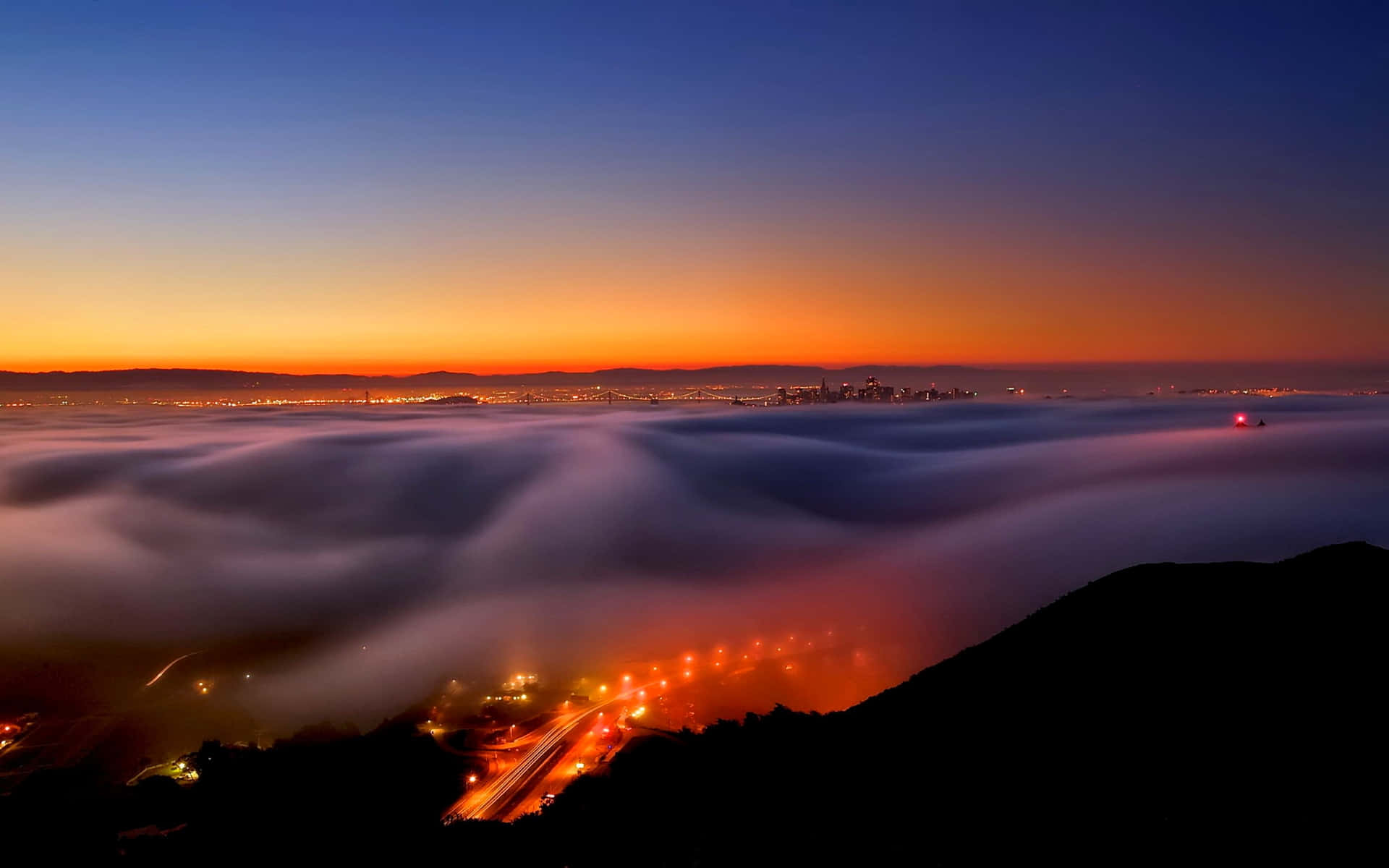 Fog 桌布 壁紙 — 城市天際線在黎明時從一片發光的霧海中浮現，下方蜿蜒的高速公路帶有光線條紋。