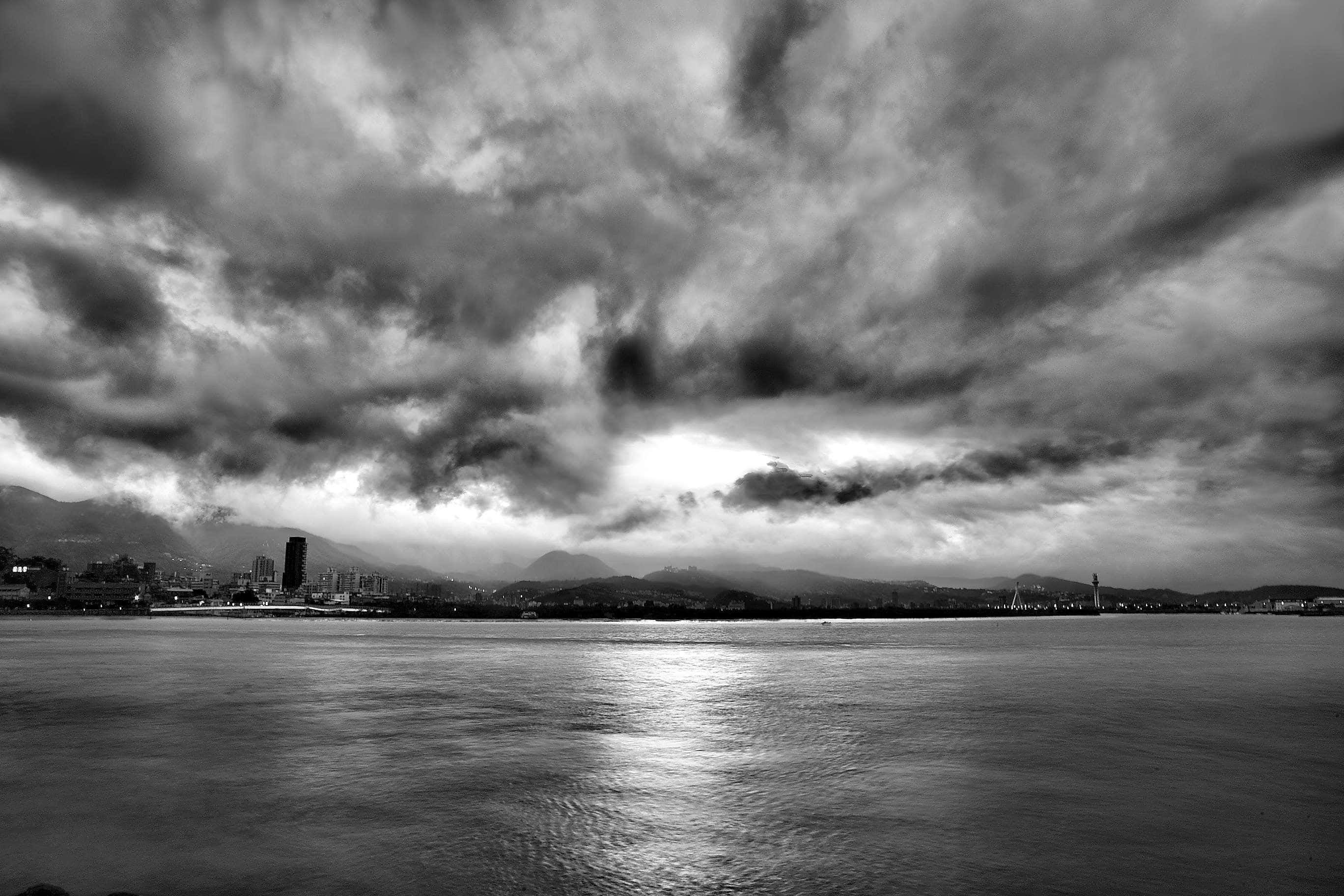 Black And White Sky 桌布 壁紙 — 城市天際線橫跨水面的戲劇性黑白景觀，陰沉的暴風雲下光線穿透。
