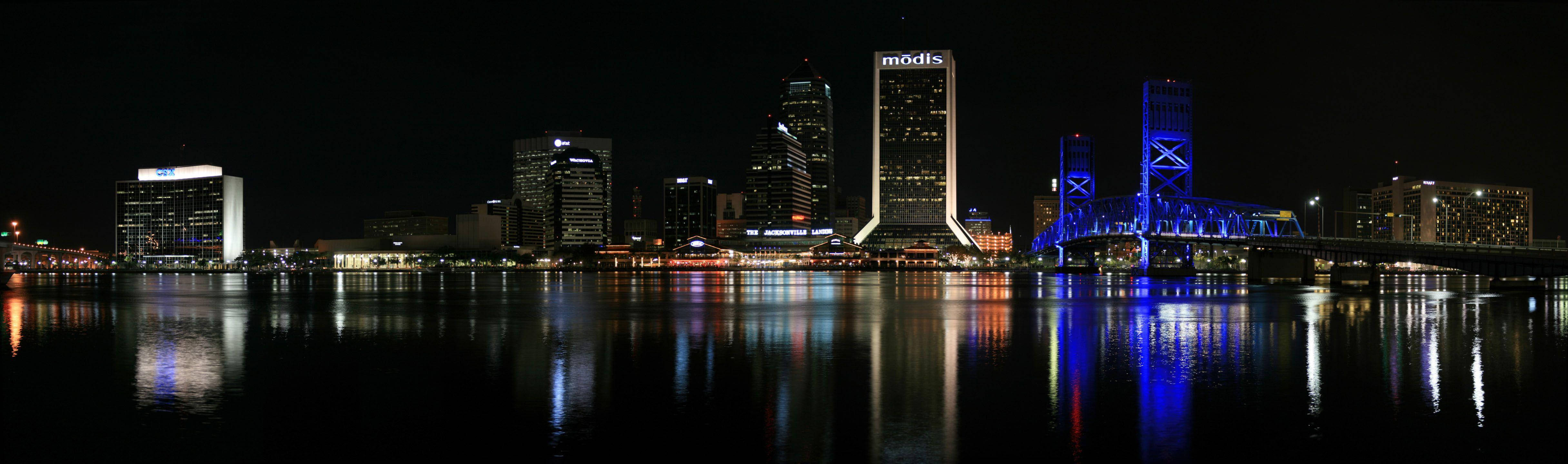 Jacksonville 桌布 壁紙 — 城市天際線的夜間全景，有燈火通明的摩天大樓和發光的藍色橋樑倒映在平靜的水面上。