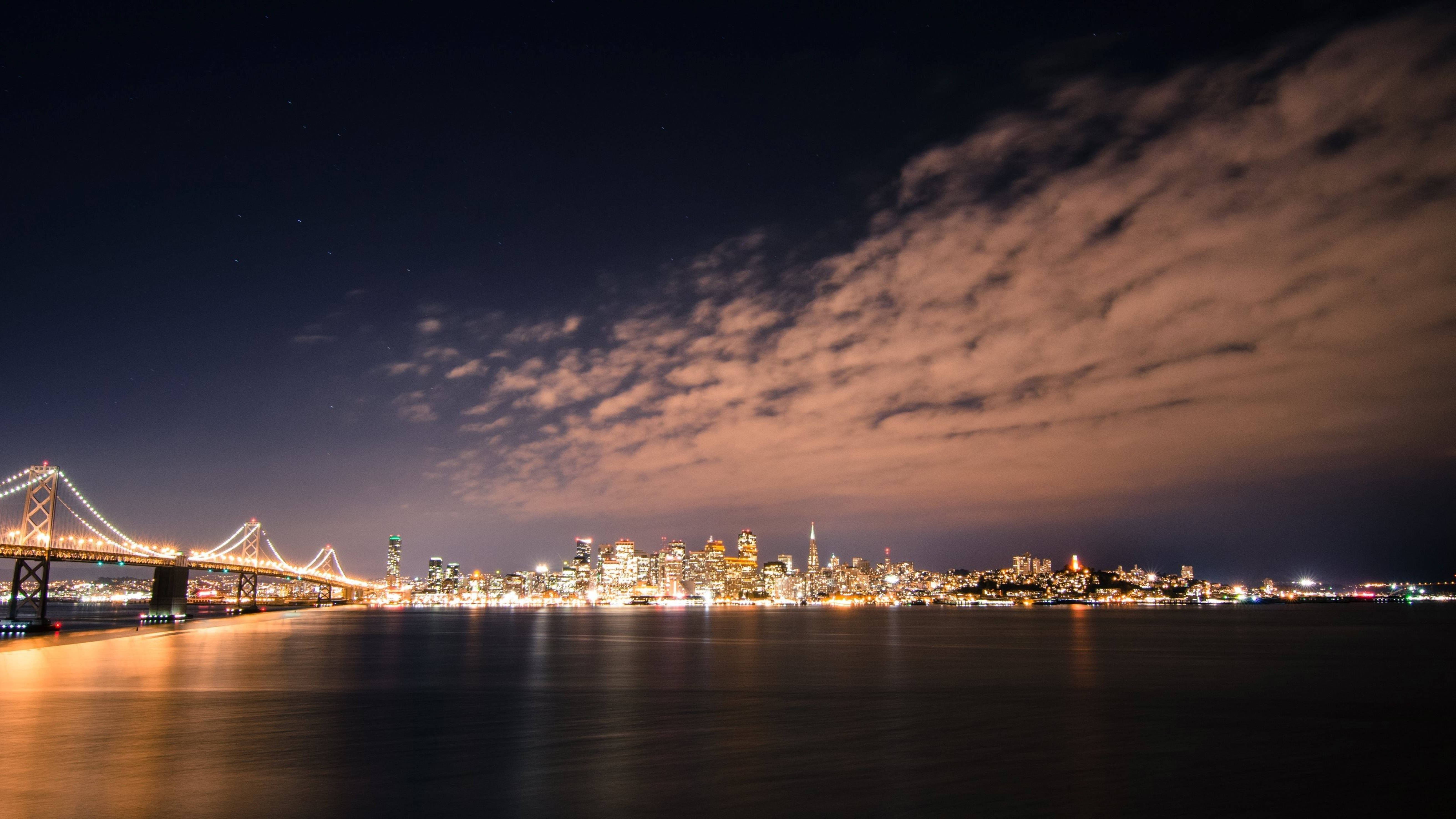 風景動畫 桌布 壁紙 — 城市天際線的迷人夜景，一座亮燈的橋樑在平靜的水面上倒映，星空下雲朵閃爍。