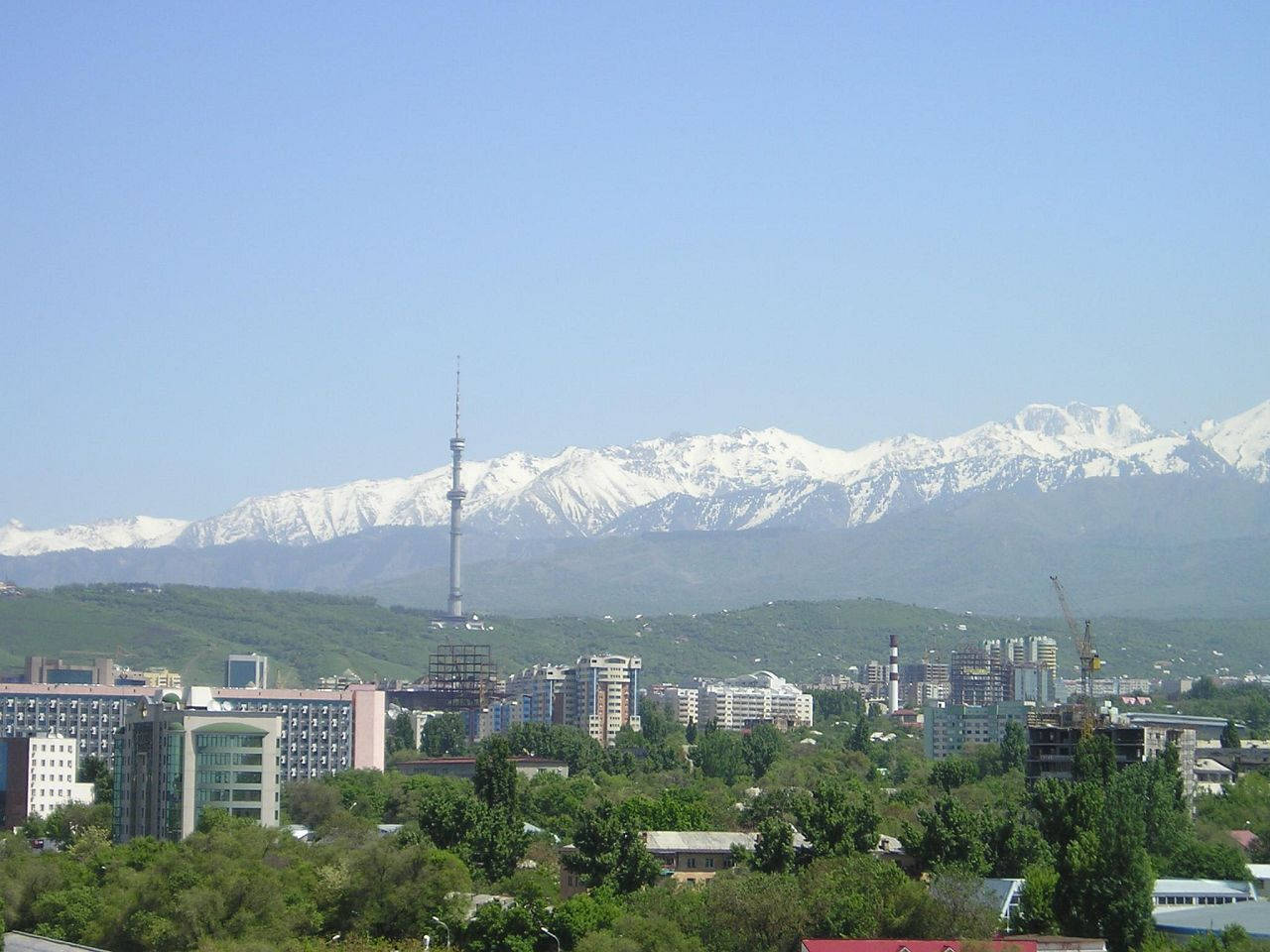 Kazakhstan 桌布 壁紙 — 城市景觀，現代建築和高塔坐落在鬱鬱蔥蔥的綠色山丘和白雪覆蓋的山脈之間，天空清澈湛藍。