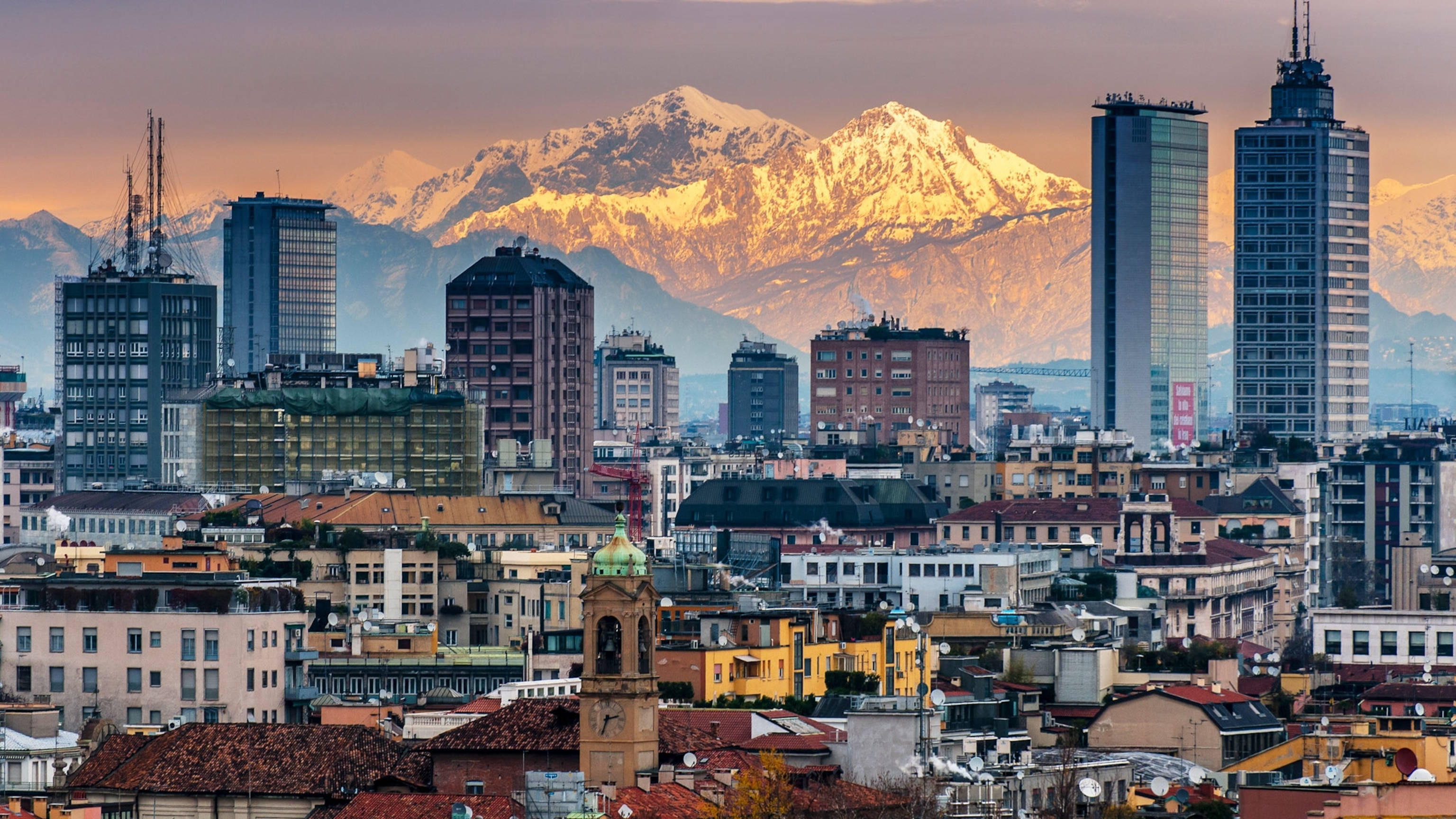 Milan 桌布 壁紙 — 城市景觀，現代摩天大樓與歷史建築以夕陽餘暉中閃耀的雪山為背景。