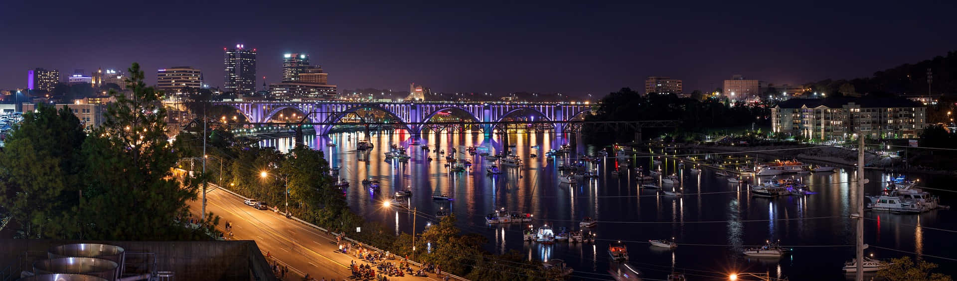 夜空 桌布 壁紙 — 城市景觀的 panoramic 夜景，一條燈火通明的橋樑橫跨河流，河上停泊著船隻，倒影閃爍。