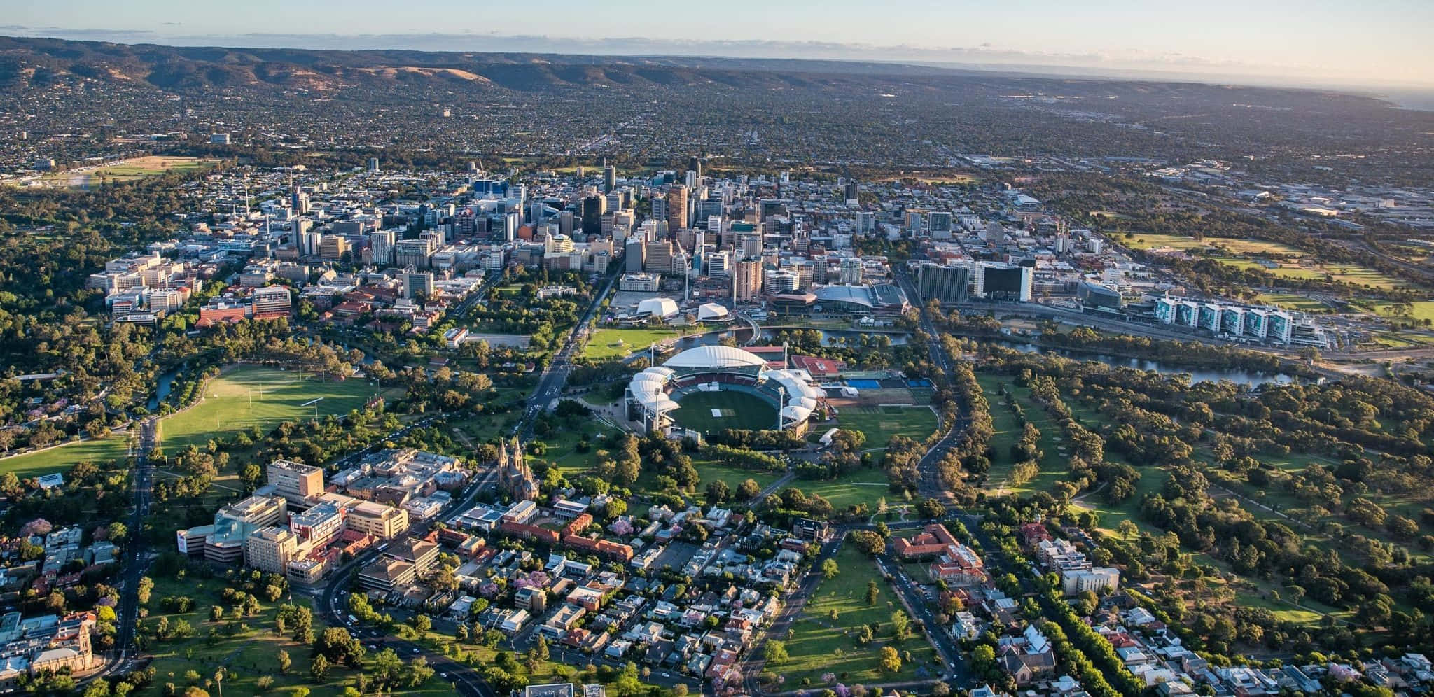 Adelaide 桌布 壁紙 — 城市鳥瞰圖，展現現代建築、綠色公園和一個在金色晨光中沐浴的大型體育場。