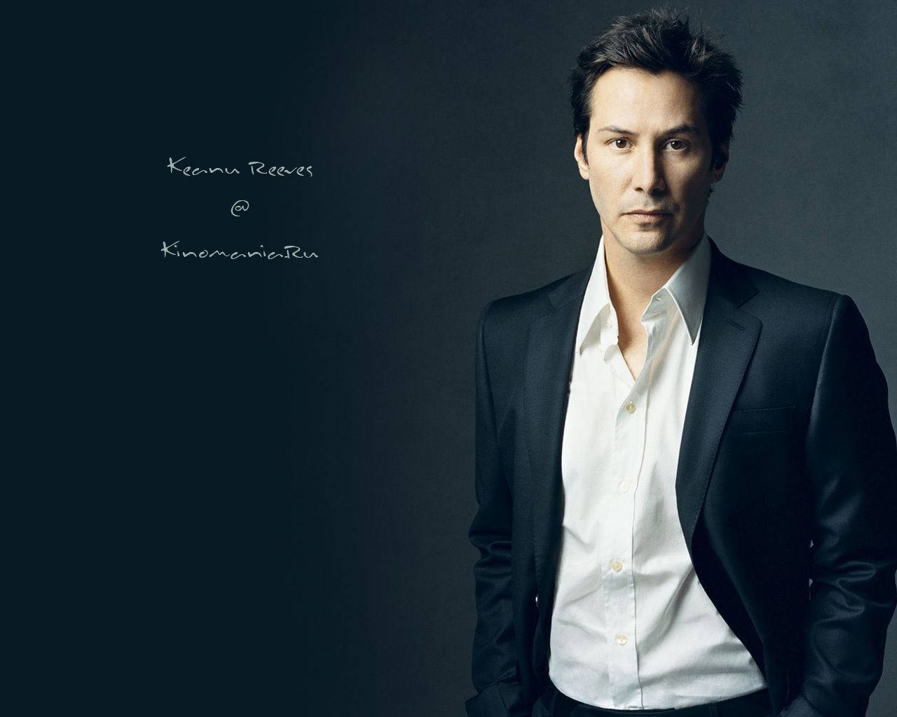 Keanu 桌布 壁紙 — 基努·李維自信地站在深色西裝和白色襯衫中，背景是陰鬱的灰色，柔和的光線突顯他強烈的目光。