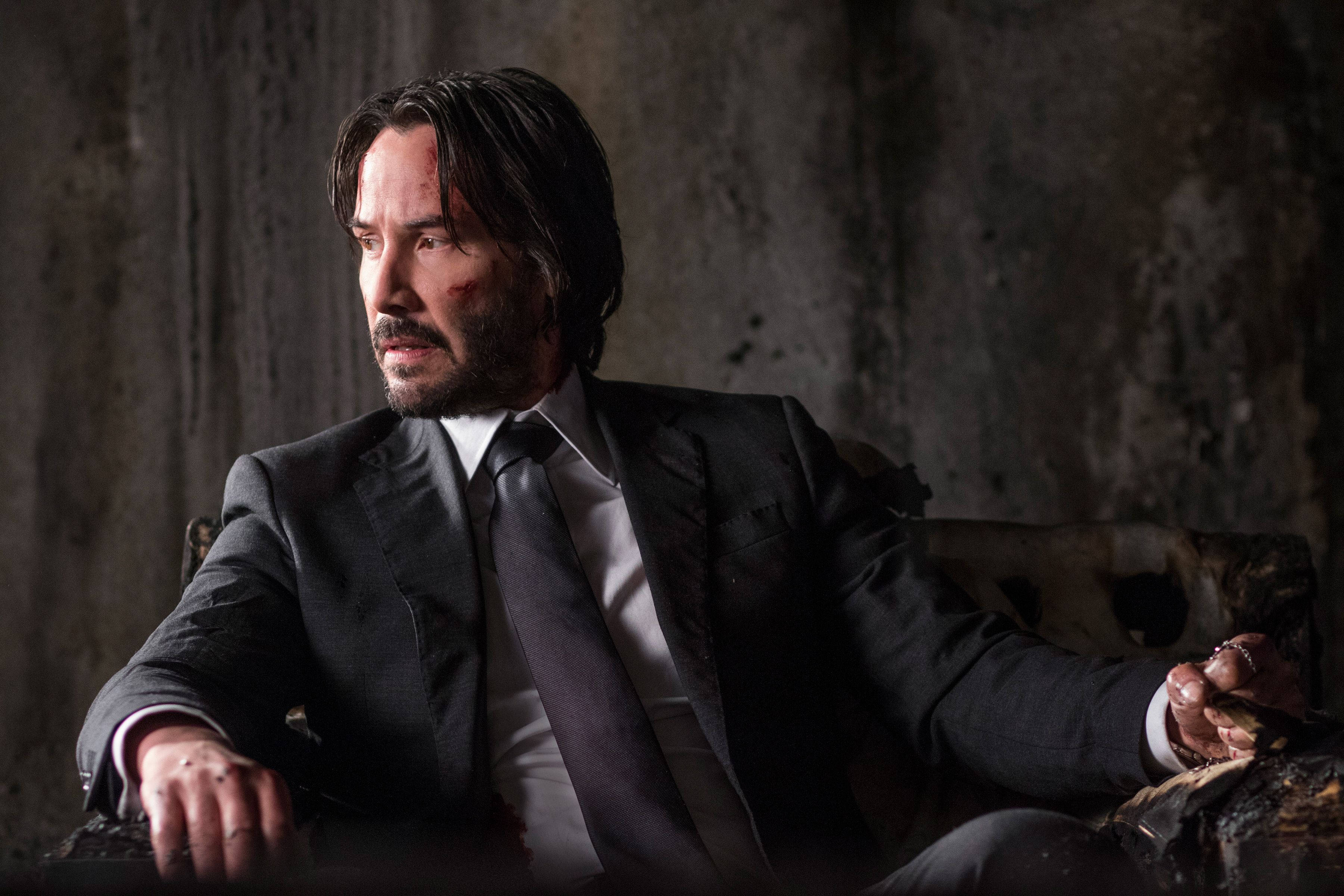 John Wick 桌布 壁紙 — 基努·李維飾演約翰·維克，坐在一張破舊的椅子上，靠著骯髒的混凝土牆，滿臉鮮血，眼神凌厲。