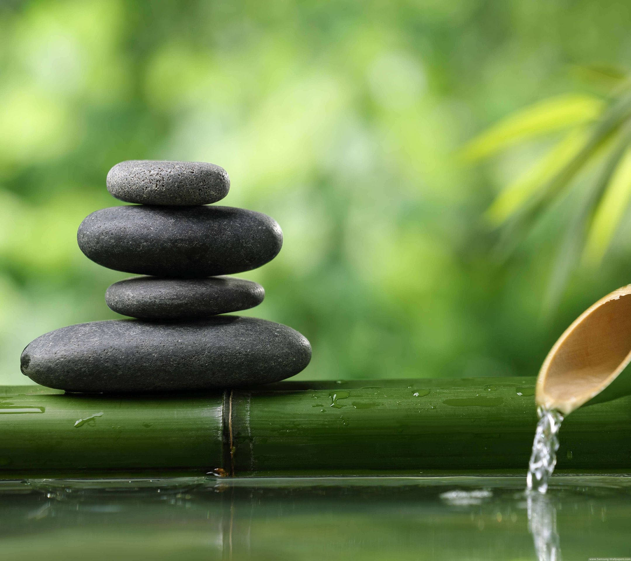 禪 桌布 壁紙 — 堆疊的平滑灰色石頭放在竹木上，旁邊有流水，在寧靜的綠色花園中。