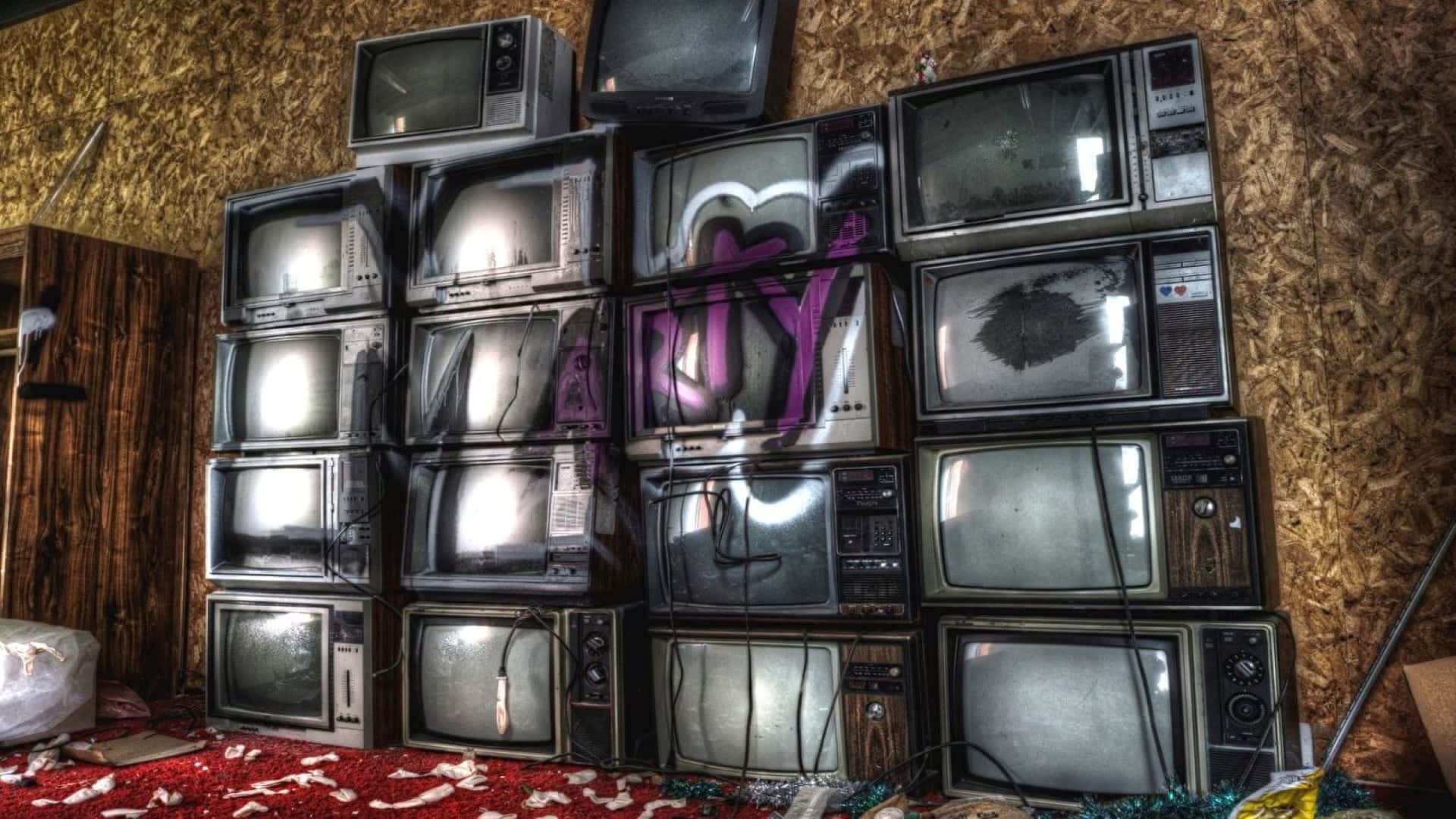 Television Set 桌布 壁紙 — 堆疊的復古電視機，帶有塗鴉和破損螢幕，位於雜亂的房間內，有裸露的OSB牆壁和紅色地毯碎屑。