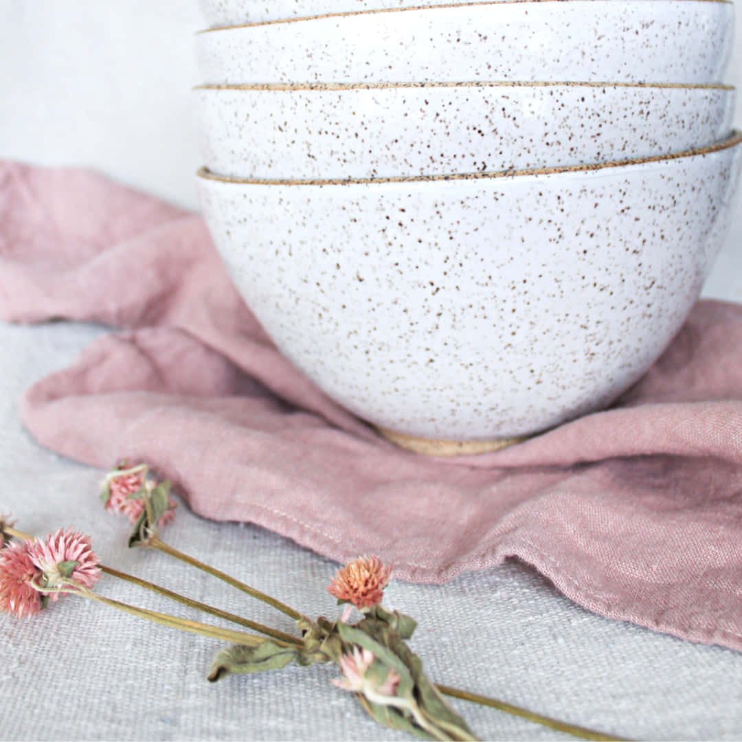 Linen 桌布 壁紙 — 堆疊的斑點陶瓷碗放在柔軟的粉紅色亞麻布上，附近散落著乾燥的粉紅色花朵。