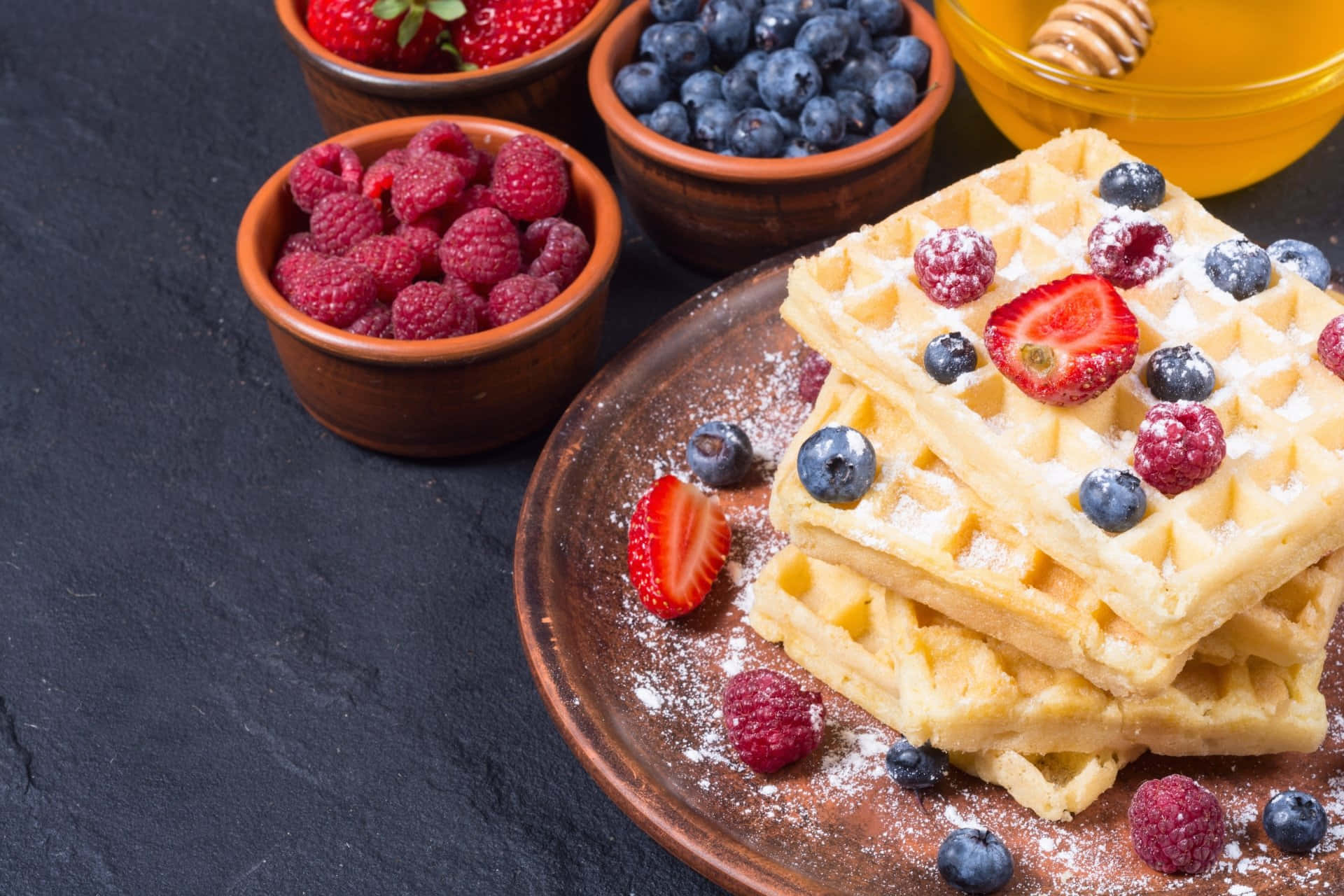 Waffle 桌布 壁紙 — 堆疊的金黃色鬆餅撒上糖粉，並放上新鮮莓果，放在鄉村風格的盤子上，旁邊有水果和蜂蜜碗。