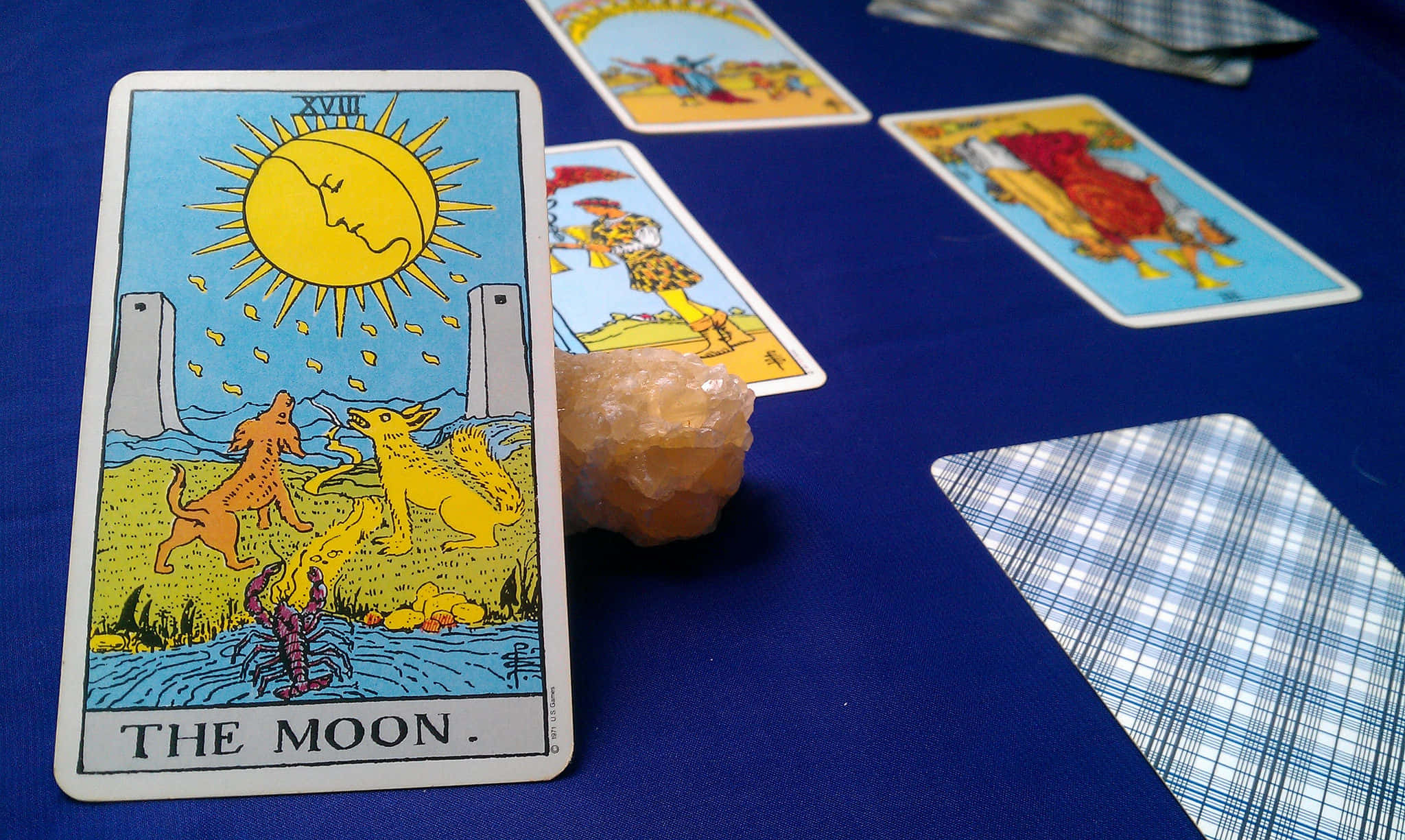 Tarot Card 桌布 壁紙 — 塔羅牌「月亮」帶有發光的太陽和嚎叫的狗，放在藍色布上，旁邊有水晶和其他牌。