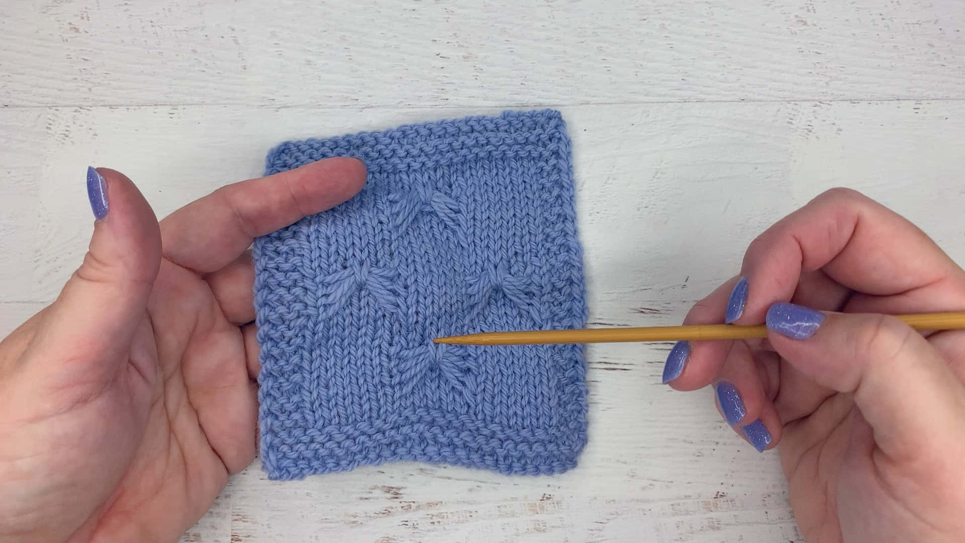 Butterfly Stitch 桌布 壁紙 — 塗有紫色指甲油的手在白色木質表面上拿著藍色編織樣本和木針。