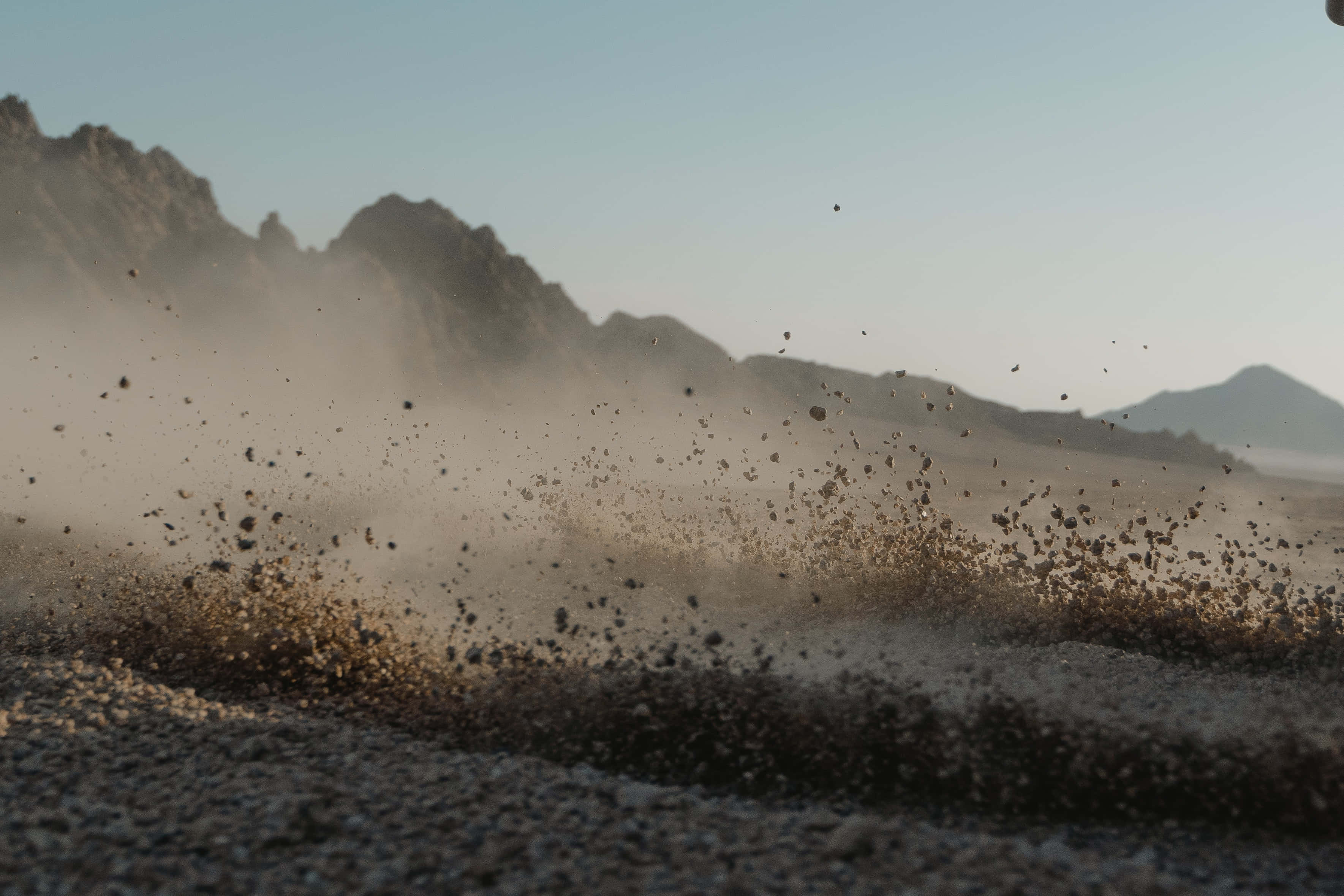 Dust 桌布 壁紙 — 塵埃與碎石在陽光照耀的沙漠地面上猛烈爆發，遠方朦朧中聳立著崎嶇的山脈。