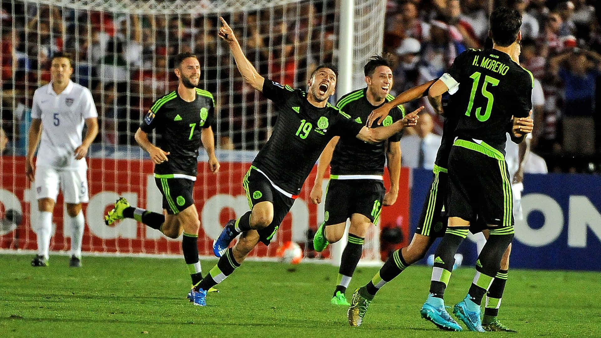 Mexico Soccer 桌布 壁紙 — 墨西哥足球運動員身穿黑色和綠色制服，在充滿活力的體育場上以狂野的動作慶祝進球。
